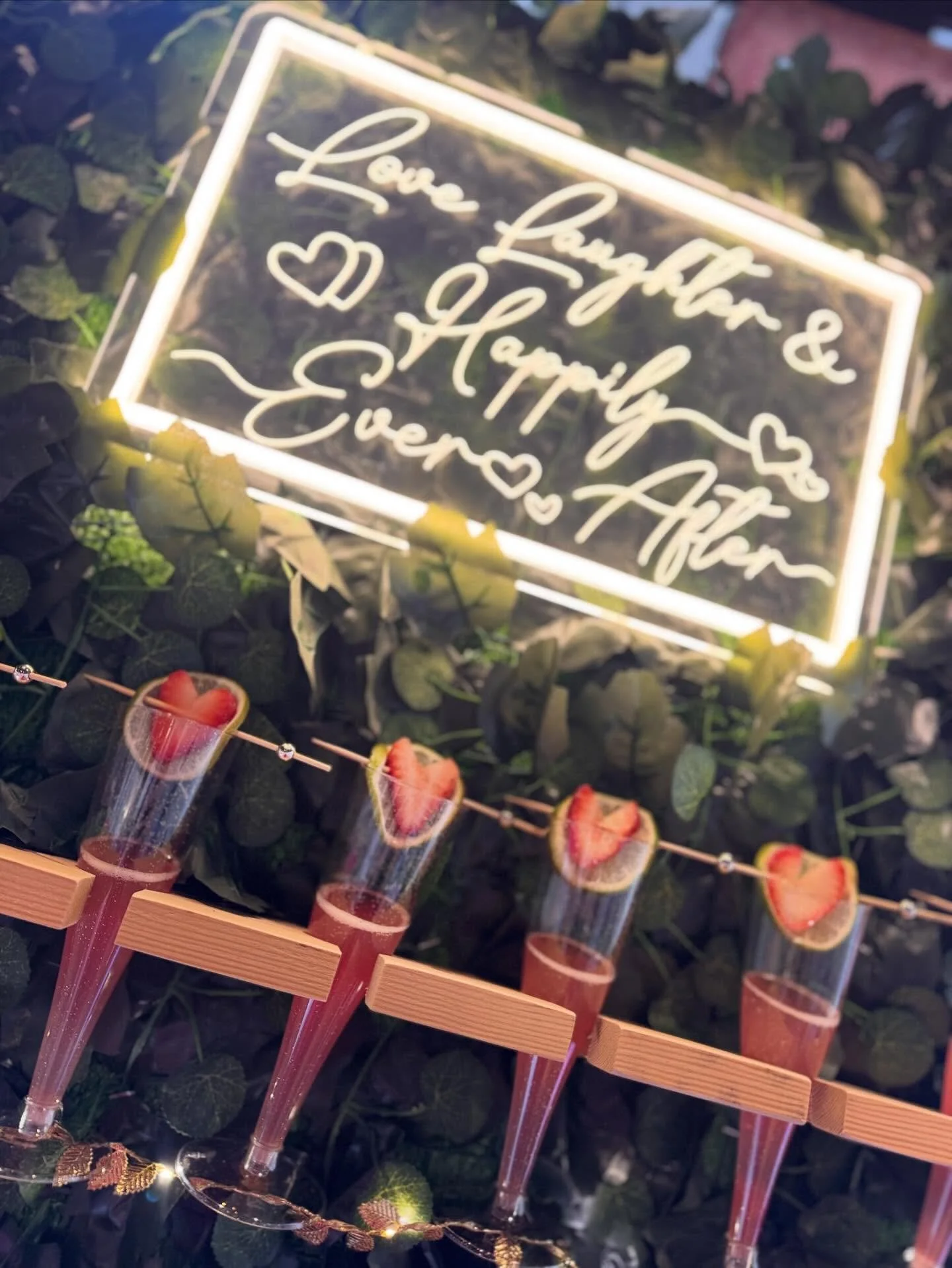 Neon sign saying 'Love Laugh & Happy' surrounded by heart shapes, underneath a string of strawberry and lemon slices attached to plastic puffs, with green leafy plants in the background.
