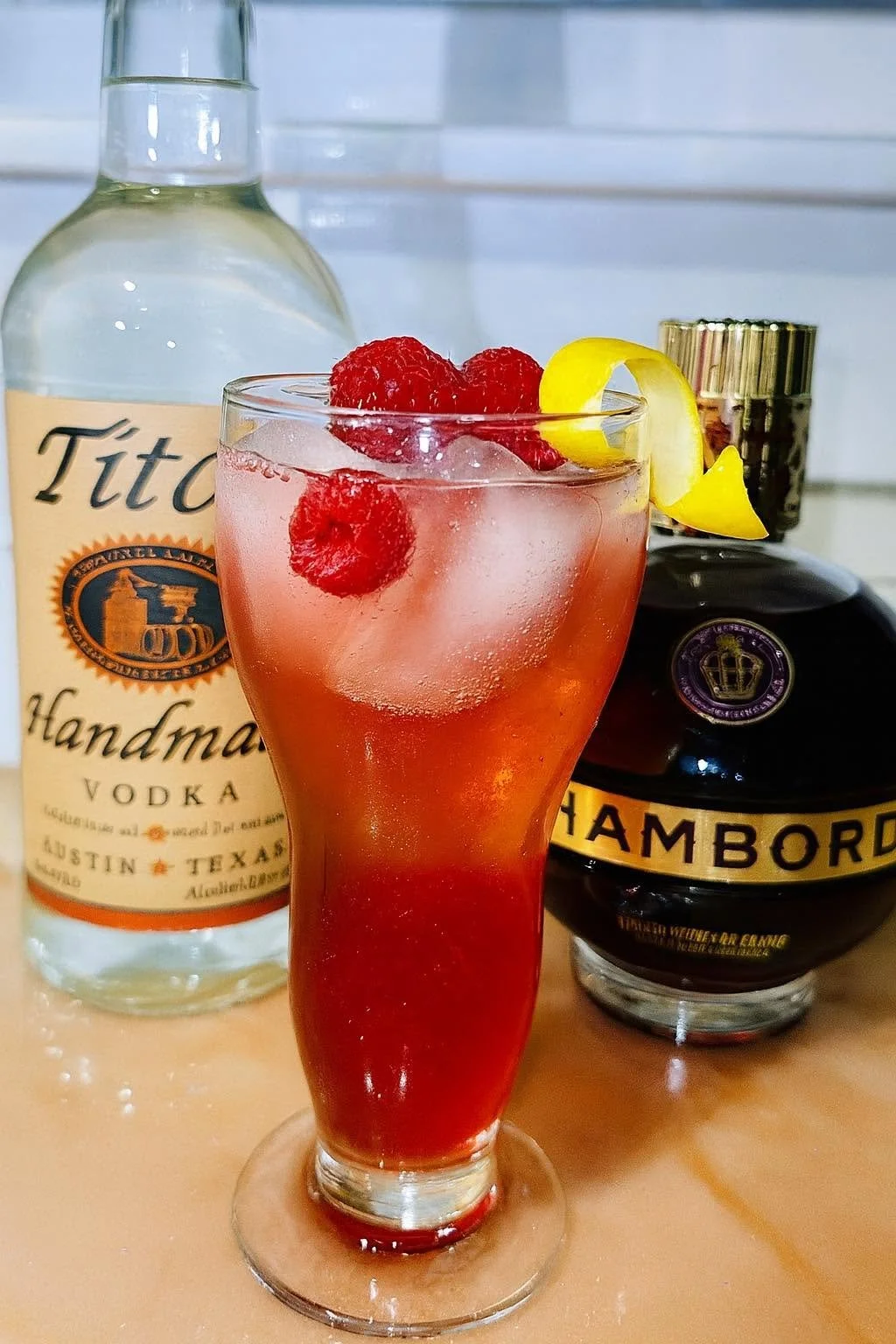 A red cocktail with raspberries and lemon slices garnished on top, placed in front of a bottle of Tito's vodka and a bottle of Chambord liqueur, on a wooden surface.