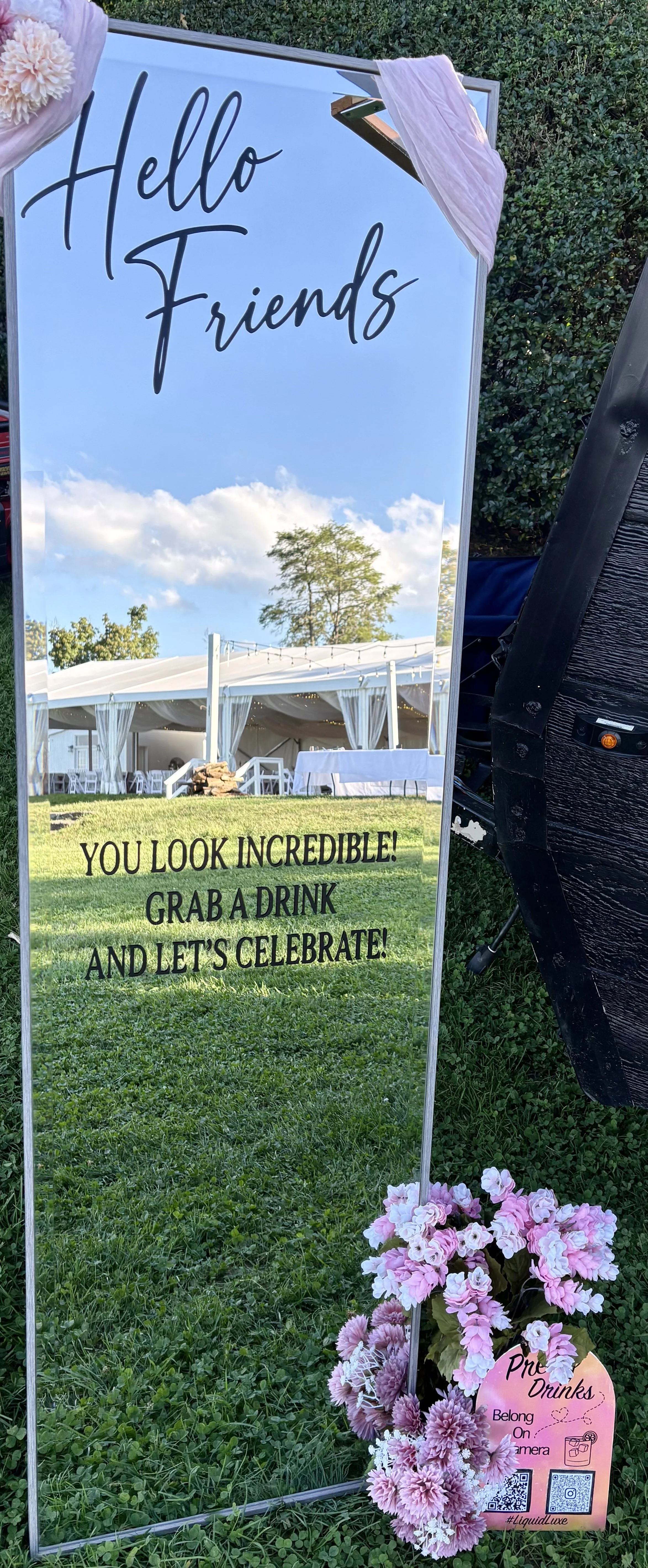 Reflective outdoor wedding or event setup sign with pink flower decorations, welcoming friends and encouraging them to grab a drink and celebrate, with a garden and marquee tent in the background.