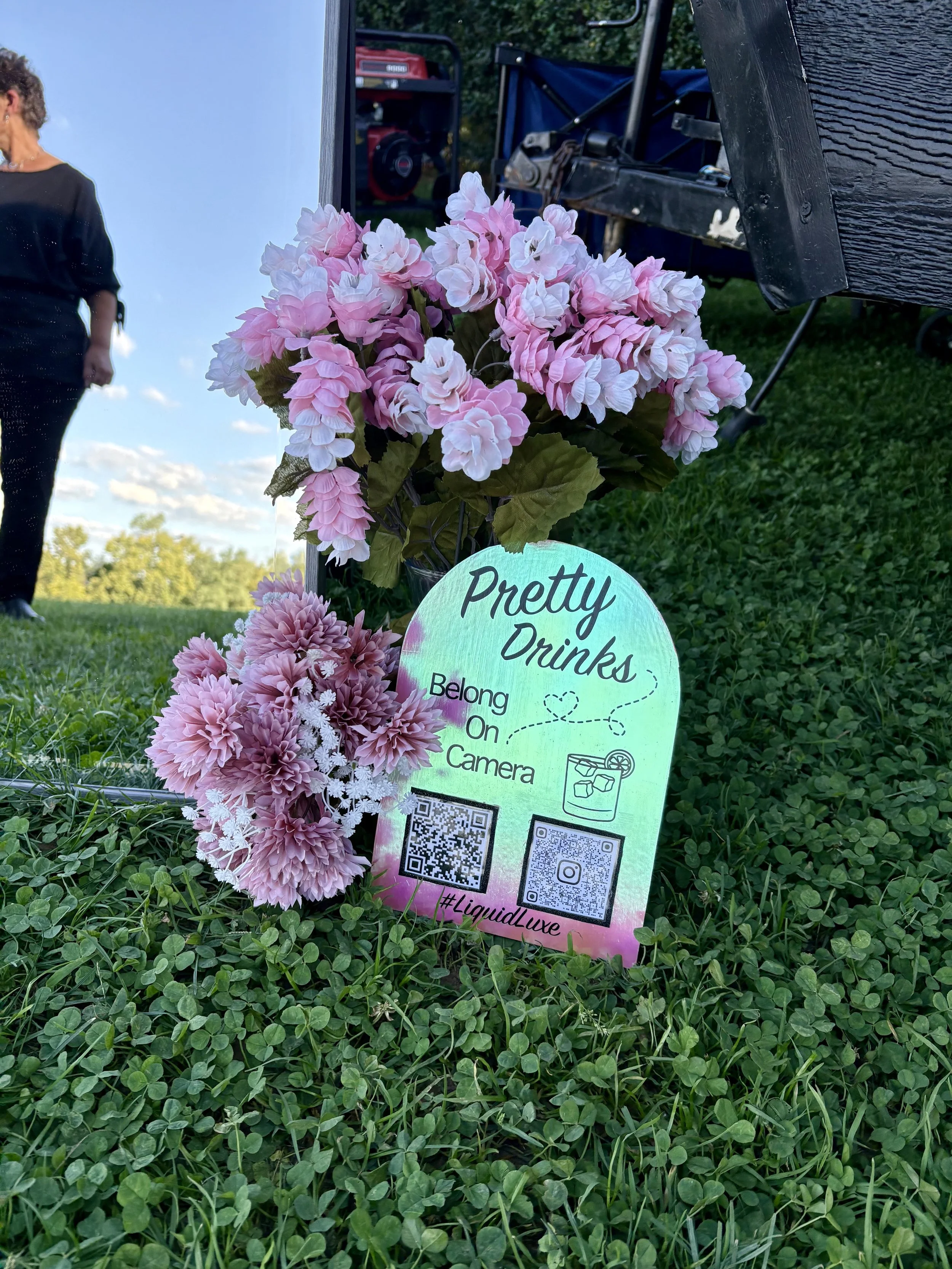 A bunch of pink and white artificial flowers placed on green grass with a signed sign that says 'Pretty Drinks' and 'Belong On Camera' along with QR codes and a hashtag #LiquidLuxe. In the background, there is a woman in black, some equipment, and a blue sky with clouds.