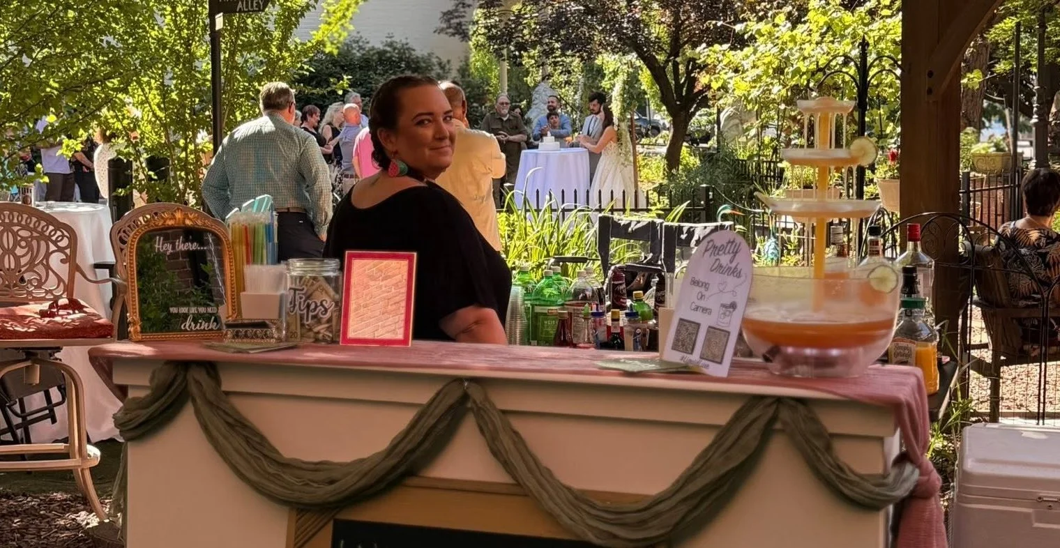 A woman standing behind a drink-serving table at an outdoor event with people socializing in the background, surrounded by trees and sunlight.