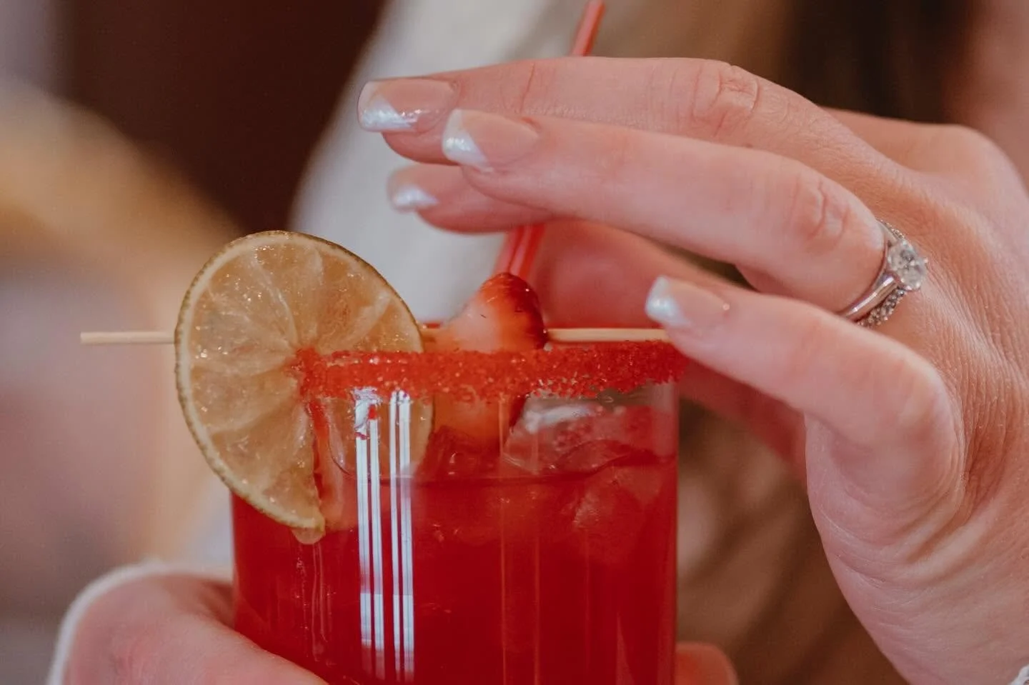 Close-up of a woman's hand holding a red cocktail with a slice of lime and a cherry garnishing the drink. The woman has manicured nails and is wearing a diamond ring.