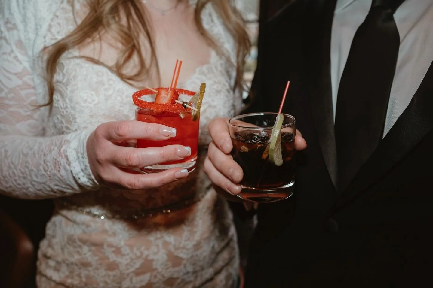 Two people holding cocktails at a celebration or party, with one person wearing a white lace dress and the other in a black suit.