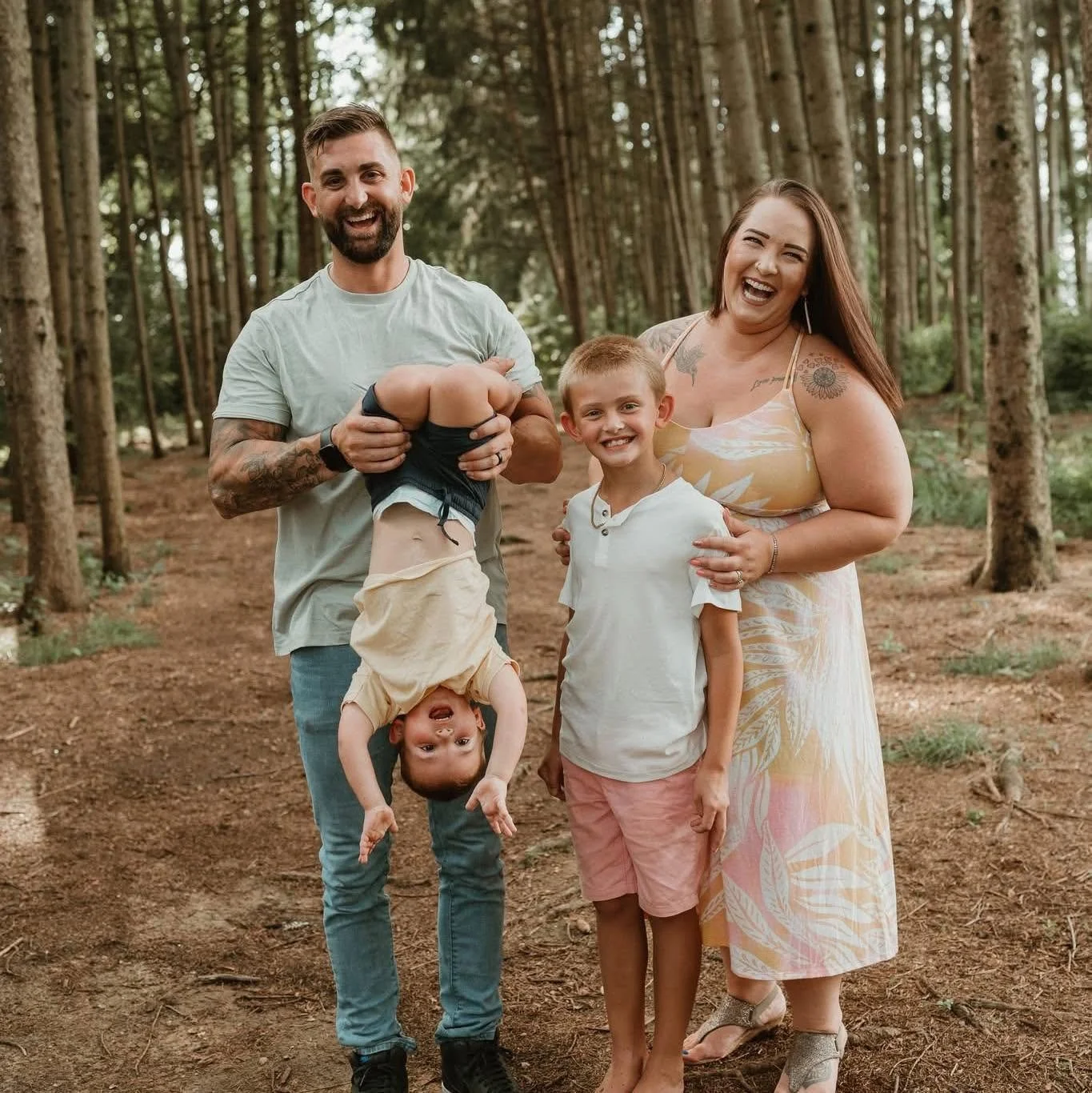 A family of five standing in a wooded forest, smiling and enjoying their time together. Two children are present, with one holding another upside down, and a woman and man are standing beside them.
