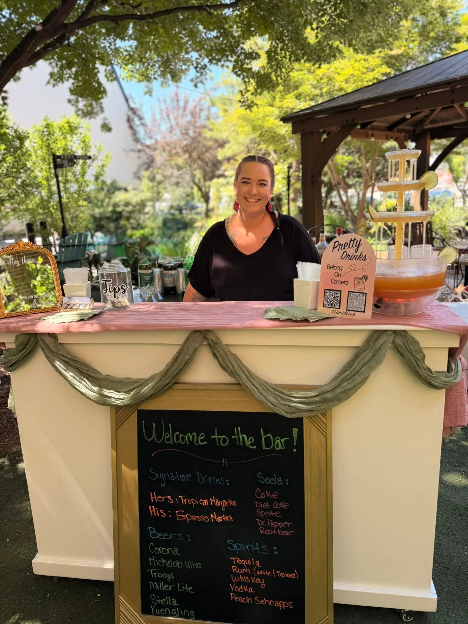 A smiling woman behind a bar made of wood and decorated with gray cloth drapes. On the bar, there are tips, a sign that says "Pretty Drinks" with a QR code, and a large drink dispenser with lemon slices. Behind her, there are trees and a gazebo in a 