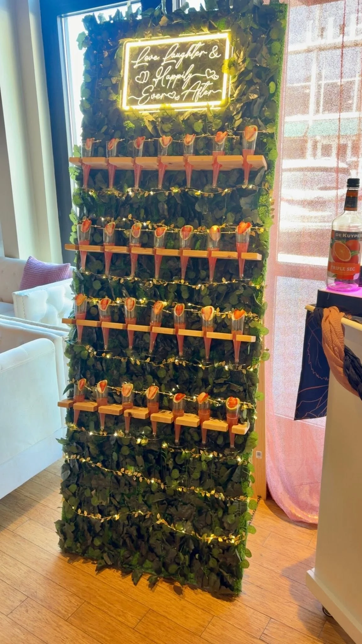 A vertical decorative display with artificial greenery and string lights, featuring a bright yellow neon sign that says 'Love, Laughter & Happiness Ever After.' Below the sign, multiple small cups filled with a pink beverage and topped with a slice of orange are arranged on wooden shelves.