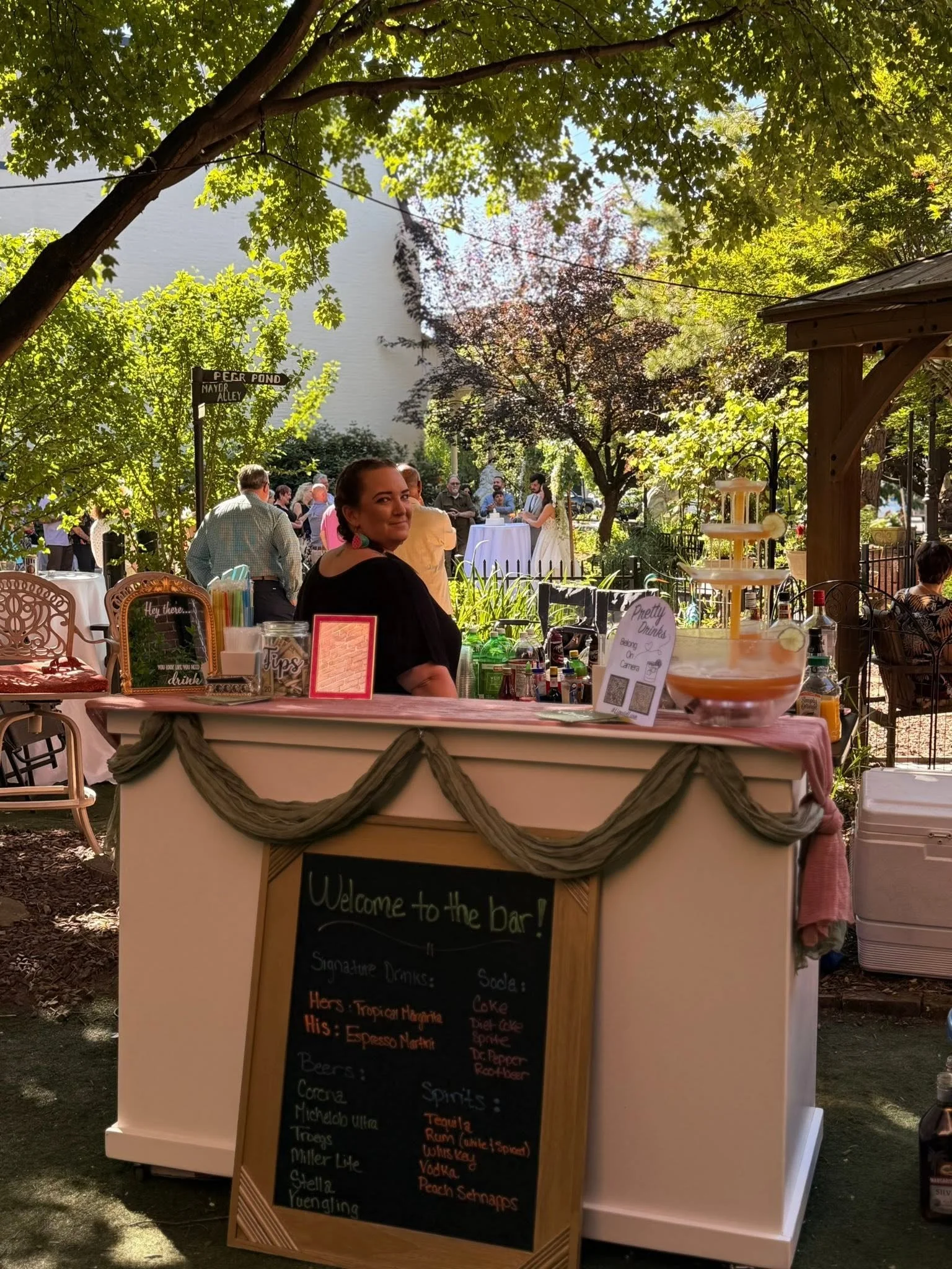 An outdoor bar with a chalkboard menu listing signature drinks and alcohol brands. A woman stands behind the bar, and people are gathered in the background under trees on a sunny day.