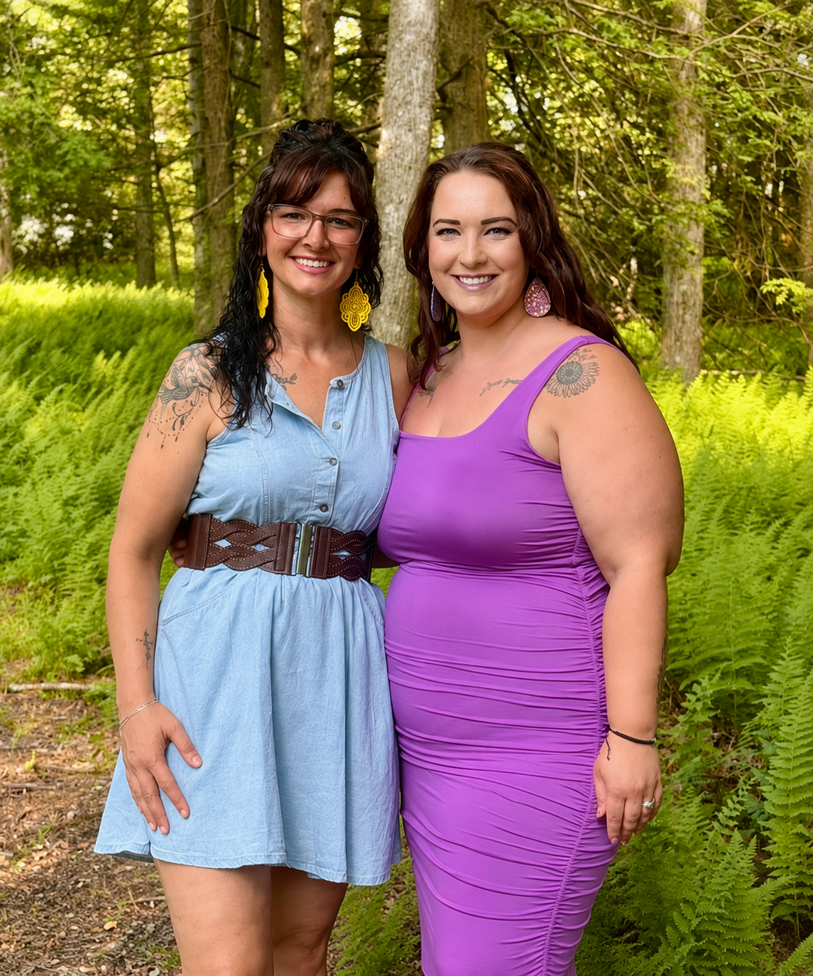 Two women standing outdoors in a wooded area, smiling for the camera. One woman is wearing a light blue dress with a brown belt and glasses, while the other is wearing a purple dress. They both have colorful earrings and visible tattoos.