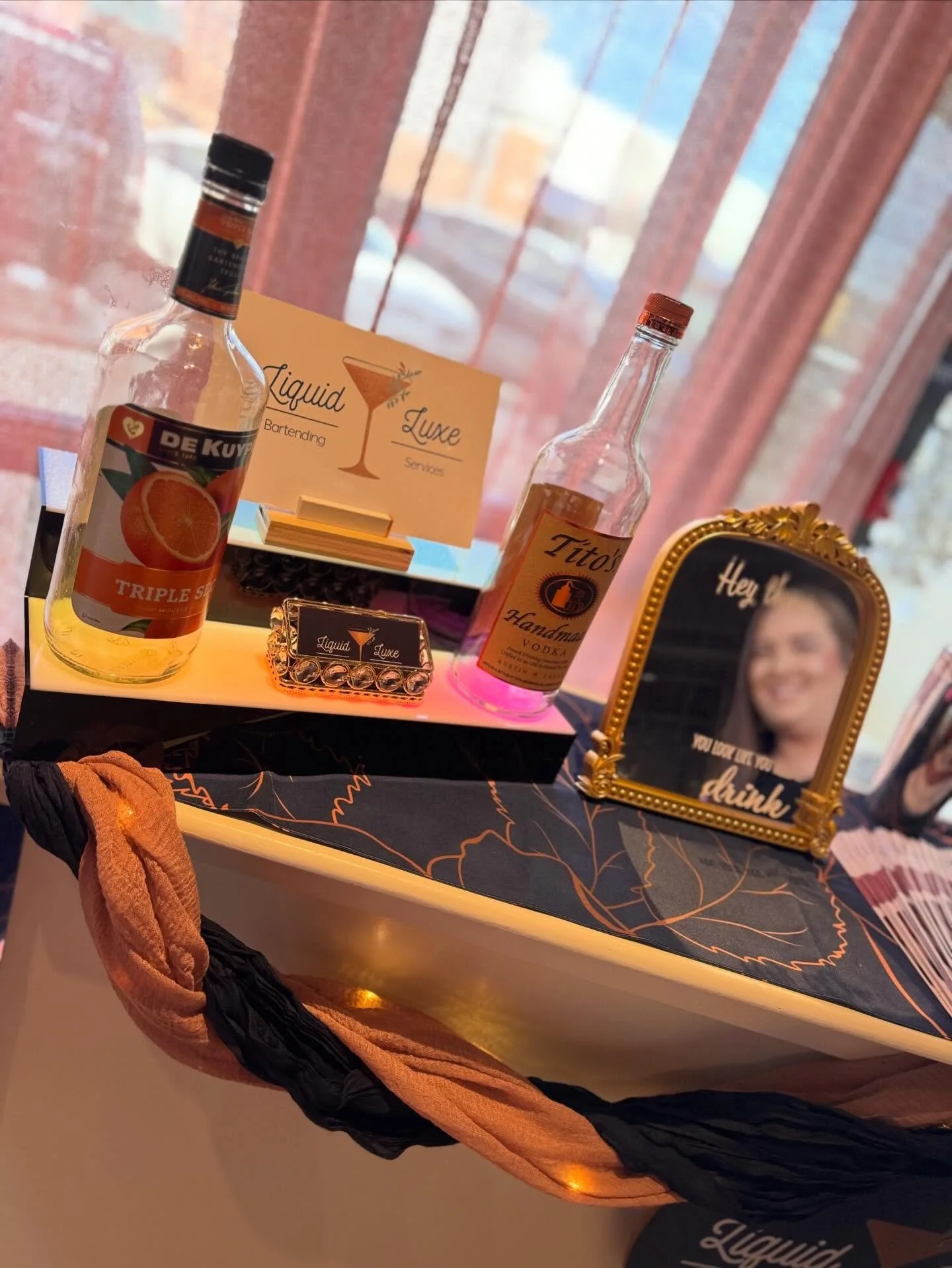 Table with bottles of liquor on display, including a triple orange flavored bottle and a vodka bottle, alongside a framed quote and a sign reading 'Liquid Luxe Bartending Services', with a woman’s face visible in the reflection of a decorative mirror