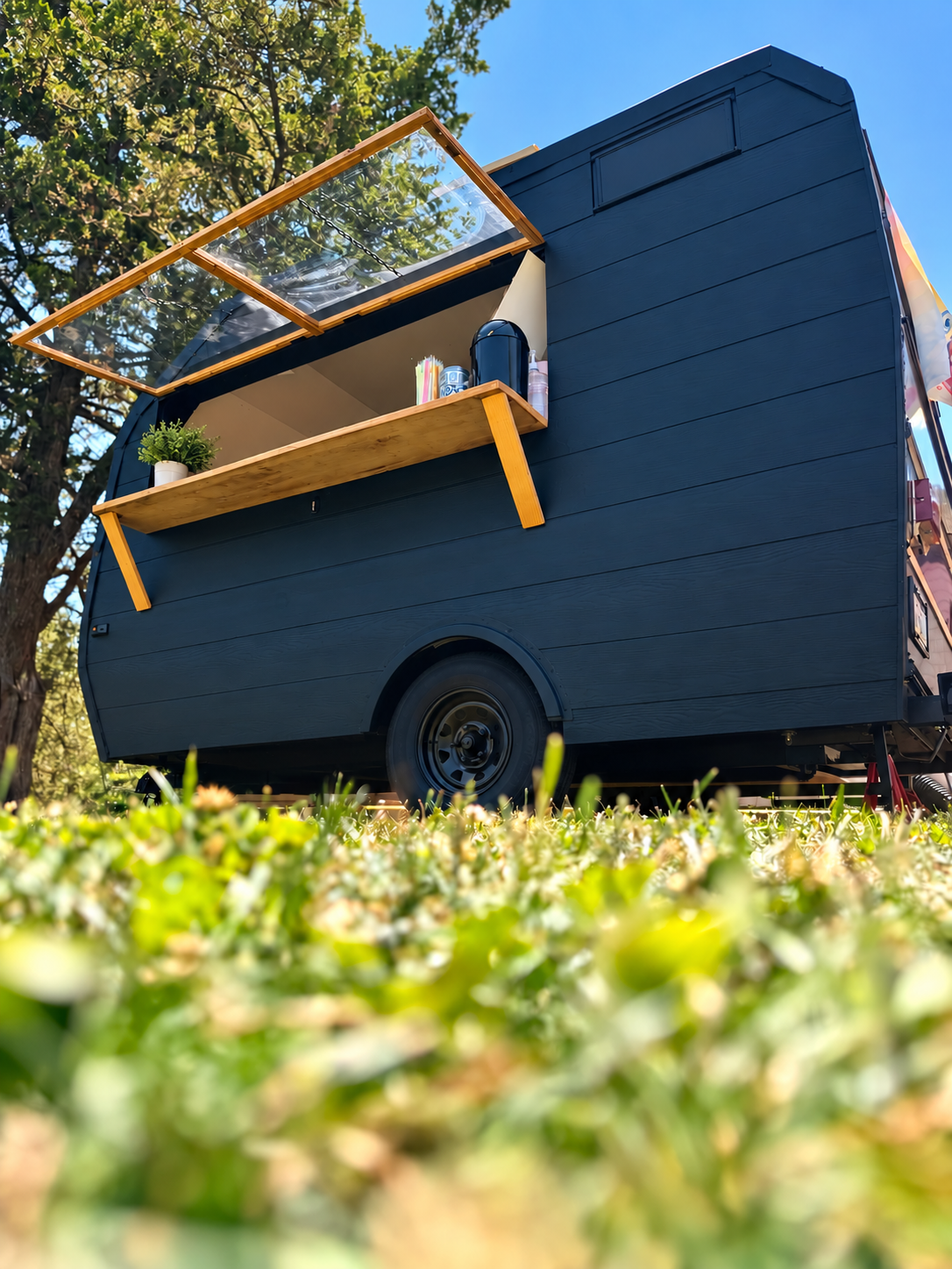 A small black camper with a wooden shelf outside, holding cups, a plant, and bottles, parked on grass with a tree and blue sky in the background.
