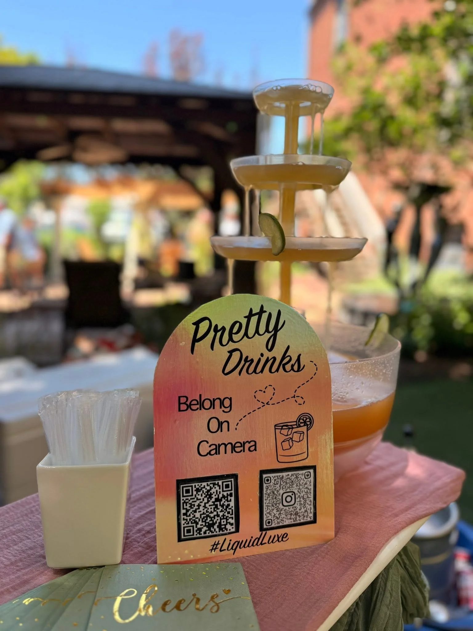 A multi-tiered drink stand with a slice of lime attached, set against a blurred outdoor background. There is a sign on the stand that reads 'Pretty Drinks' with QR codes underneath and a hashtag '#LiquidLuxe'. A small container of white straws is als