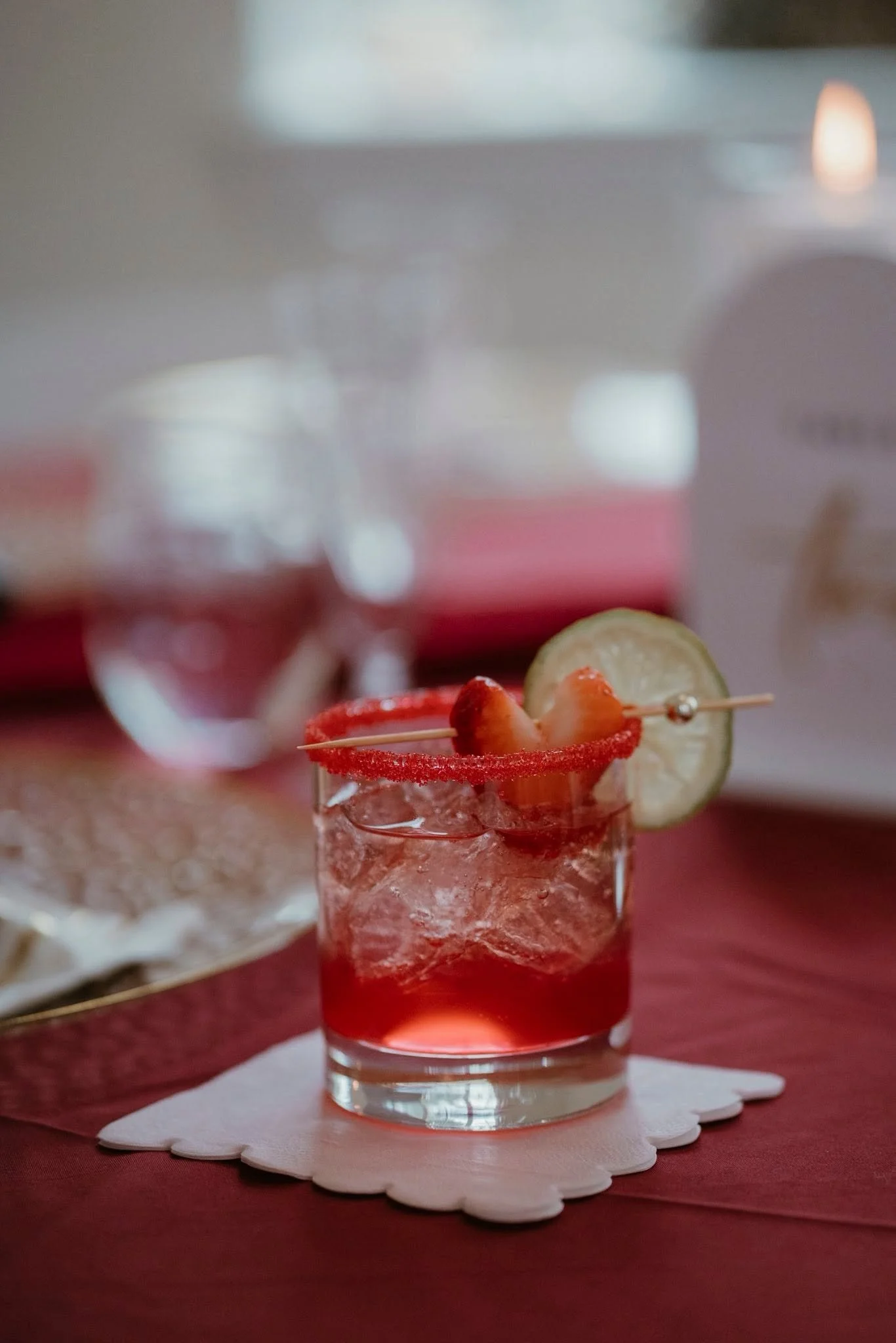 A red cocktail garnished with a lemon slice and a strawberry on a skewer, served on an ice-filled glass with a salted rim, placed on a white napkin on a red table.