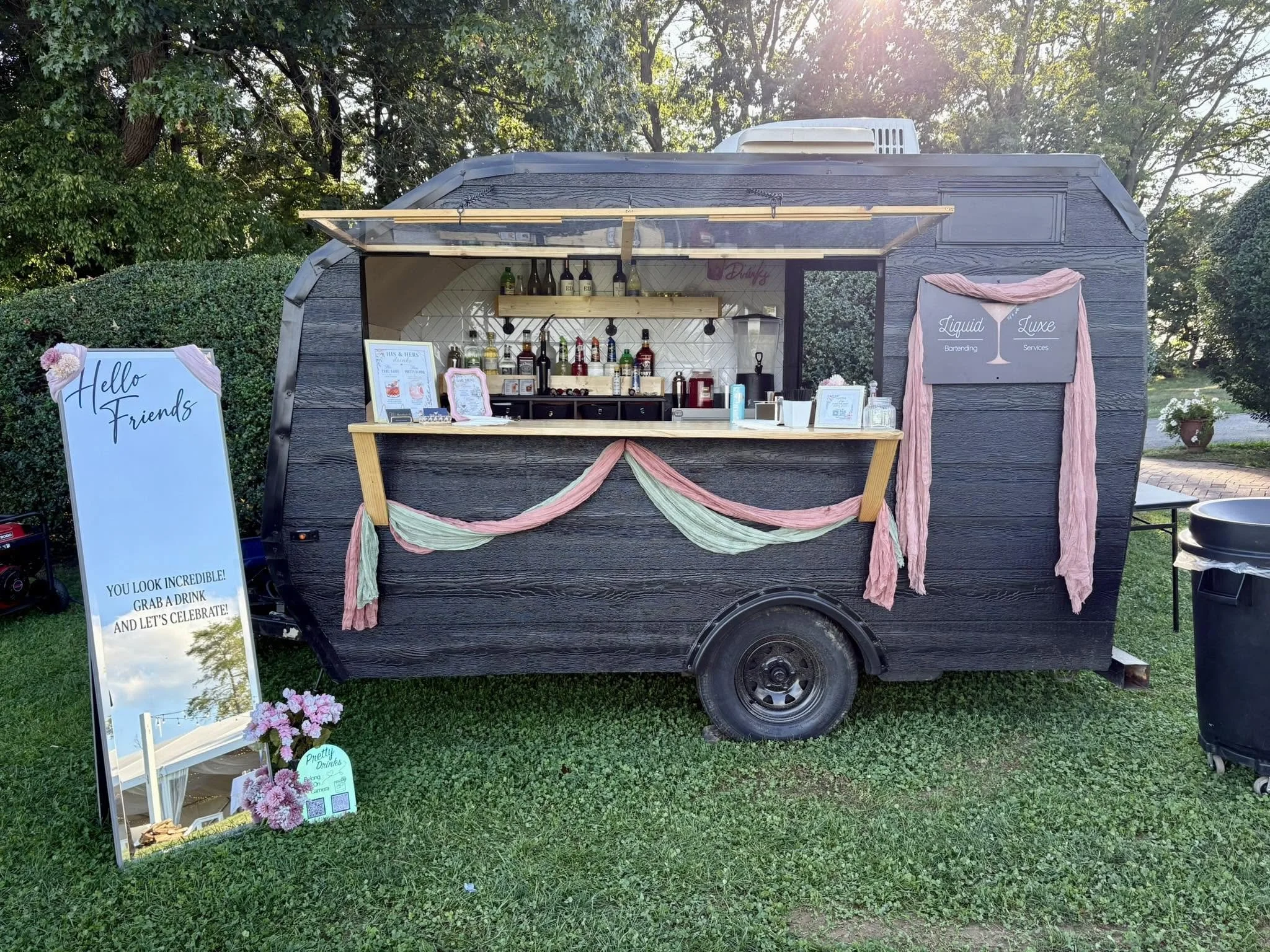 A small black mobile bar with pink and green fabric decorations, set outdoors on a grassy area. It has a window opening displaying bottles of alcohol and bar tools inside. There is a white sign with black writing on the left side that reads 'Hello Fr