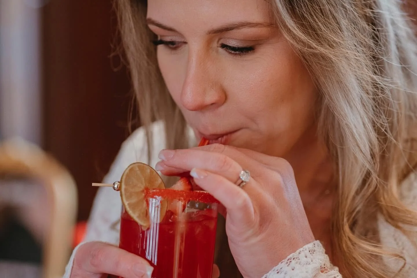 A woman drinking a red beverage through a straw, garnished with a lemon slice and a small cherry, in a glass with a decorative topper.