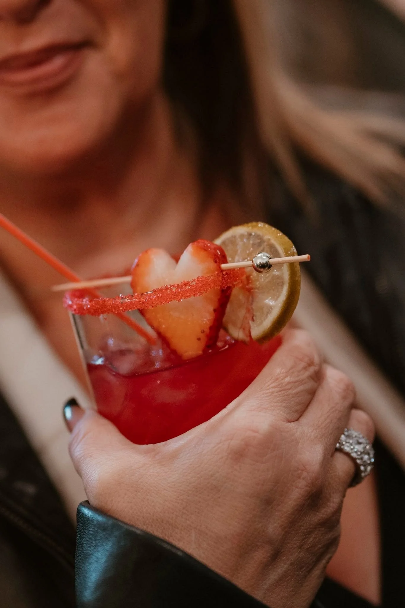 Close-up of a woman's hand holding a cocktail glass with a strawberry, lemon wedge, and red sugar rim decoration, blurred face in the background.