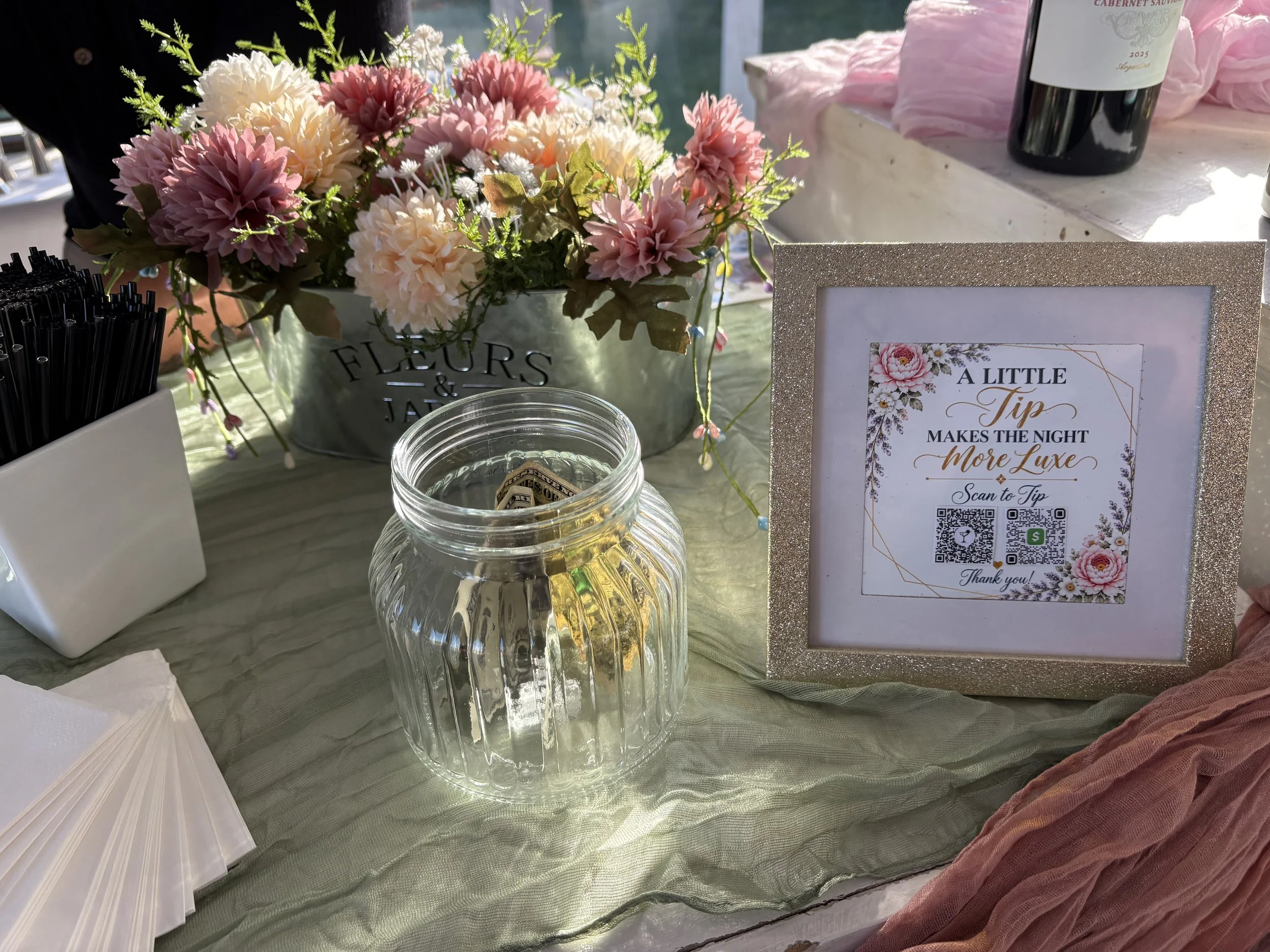 A floral centerpiece with pink and white flowers in a metal bucket, placed on a table with a green tablecloth. Nearby are black straws in a white container, a glass jar with money, pink decorations, a framed sign with a floral design, QR codes, and a
