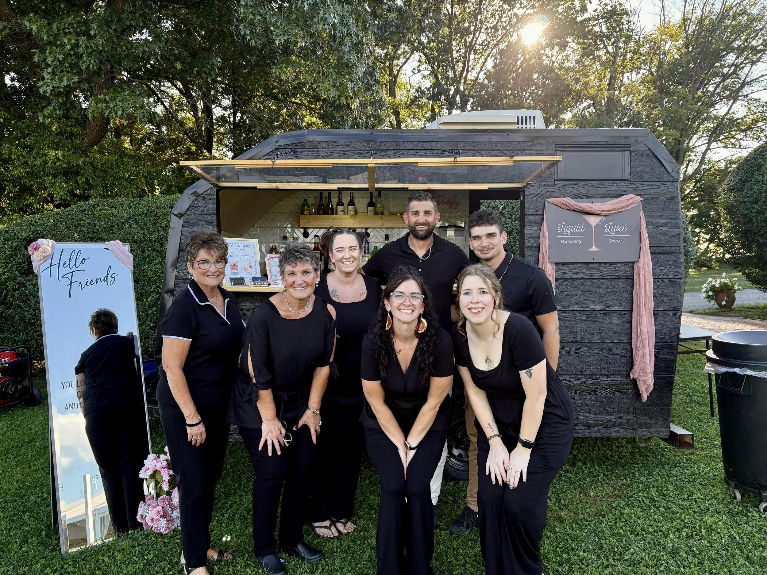 A group of nine people, mostly women, posing together outdoors in front of a black food trailer with a pink cloth and a sign that reads "Liquid Luxe". The trailer is set against a backdrop of green trees with the sun shining through.