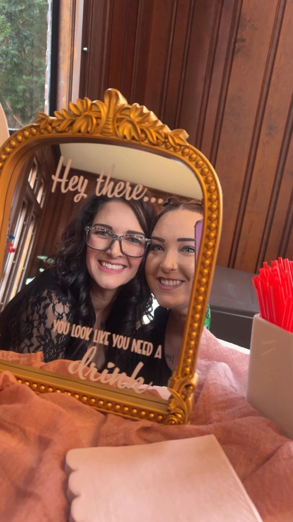 Two women smiling and taking a selfie in front of a decorative mirror with the words 'Hey there...' written on it.