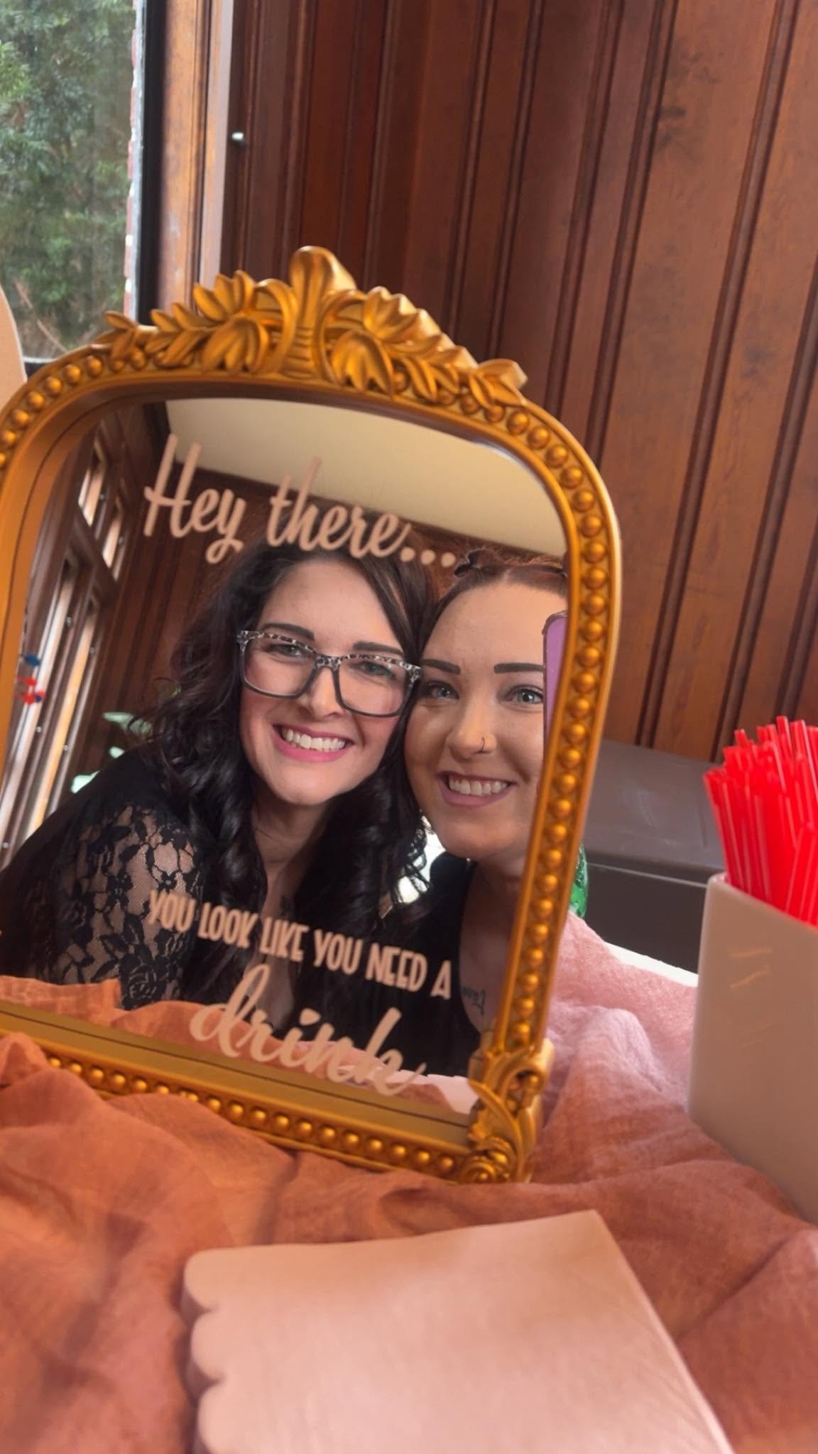 Two women smiling and taking a selfie in a mirror, with text overlay saying "Hey there..." and more text about makeup and lipstick. The mirror has a gold ornate frame, and the background shows a wood-paneled wall and a window.