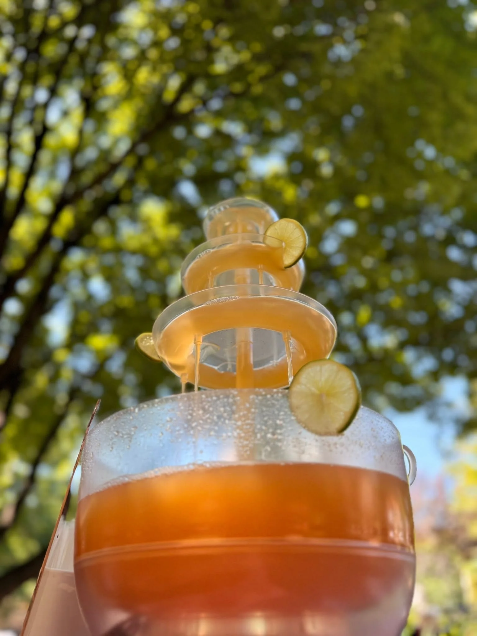 A multi-tiered orange and clear glass beverage fountain filled with an orange-colored drink, garnished with lemon slices, set outdoors with a background of green trees and sunlight.