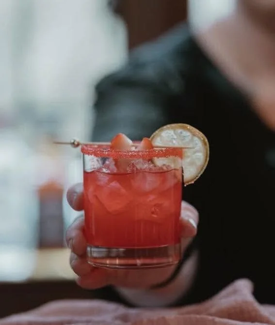 A person holding a red cocktail garnished with a lemon slice and a cherry, served with ice cubes in a short glass.