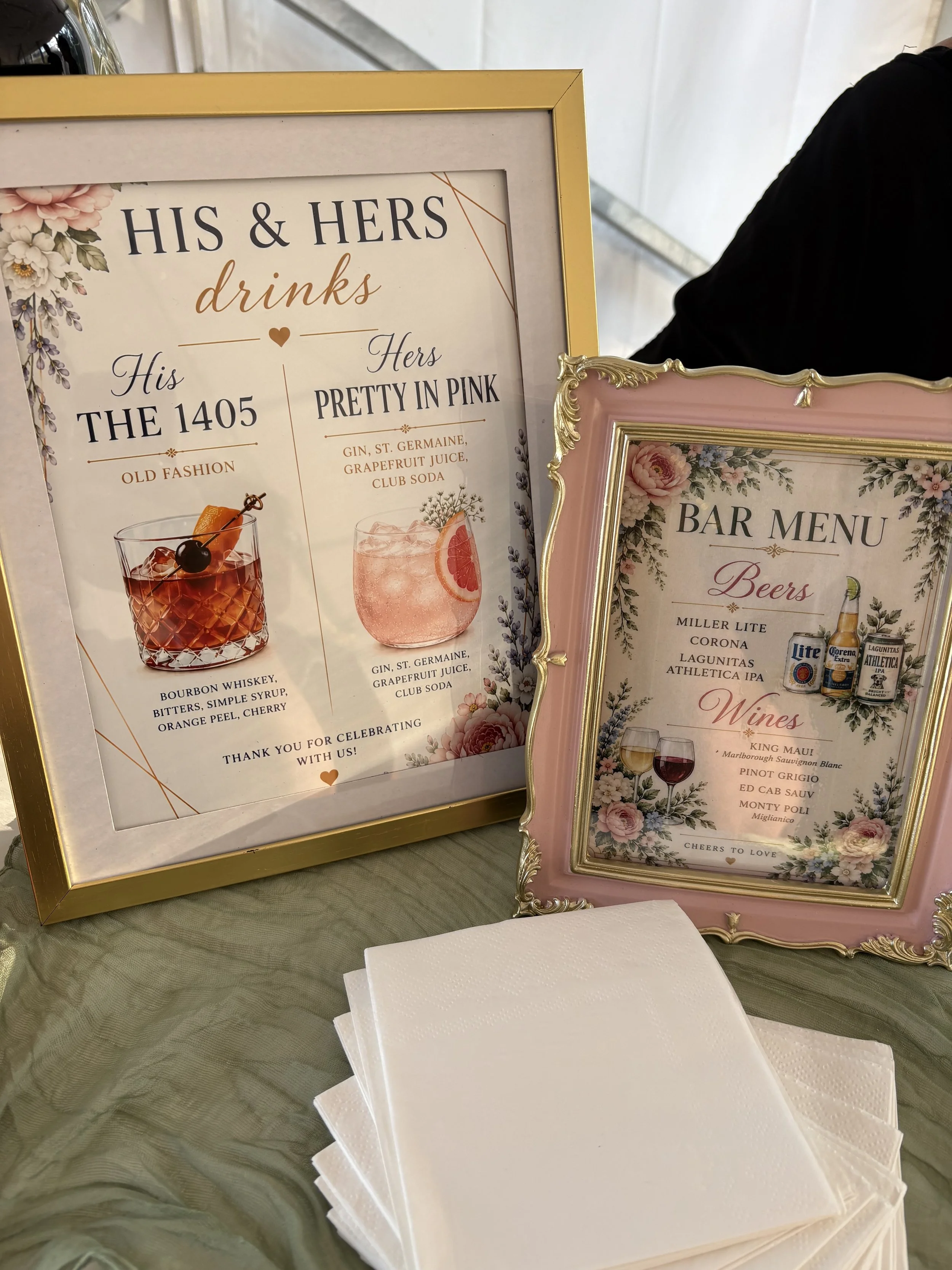 Signs displaying drink and bar menu options at an event. The first sign features cocktails with ingredients like bourbon whiskey, bitters, simple syrup, orange peel, and cherry, with a gold frame. The second sign lists wines such as King Mauf, Marlborough Sauvignon Blanc, Pinot Grigio, Ed Cab Sauv, and Monty Poli, with a pink ornate frame and floral decoration.