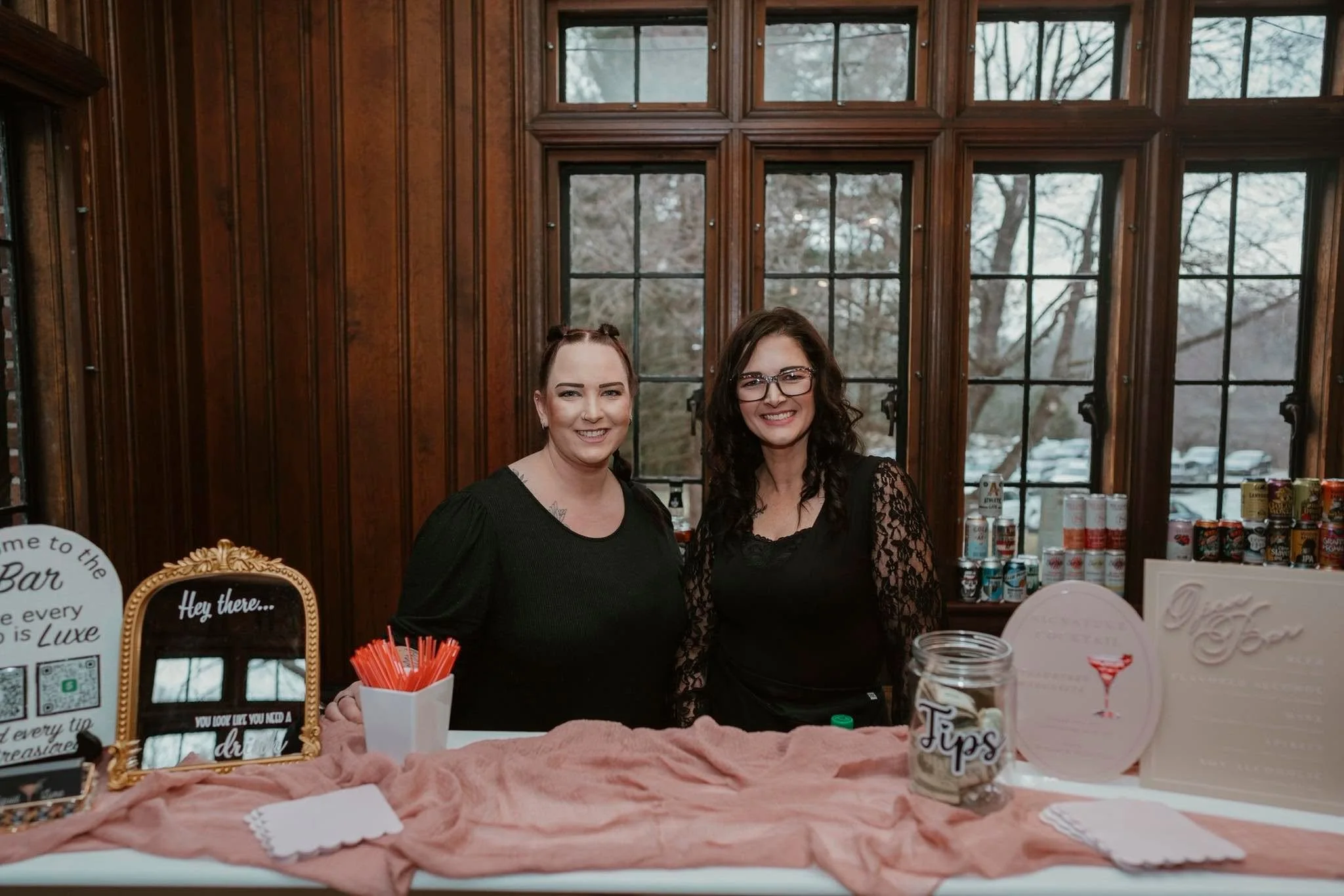 Two women stand behind a table with promotional items inside a wooden building with large window panes, greenery visible outside.