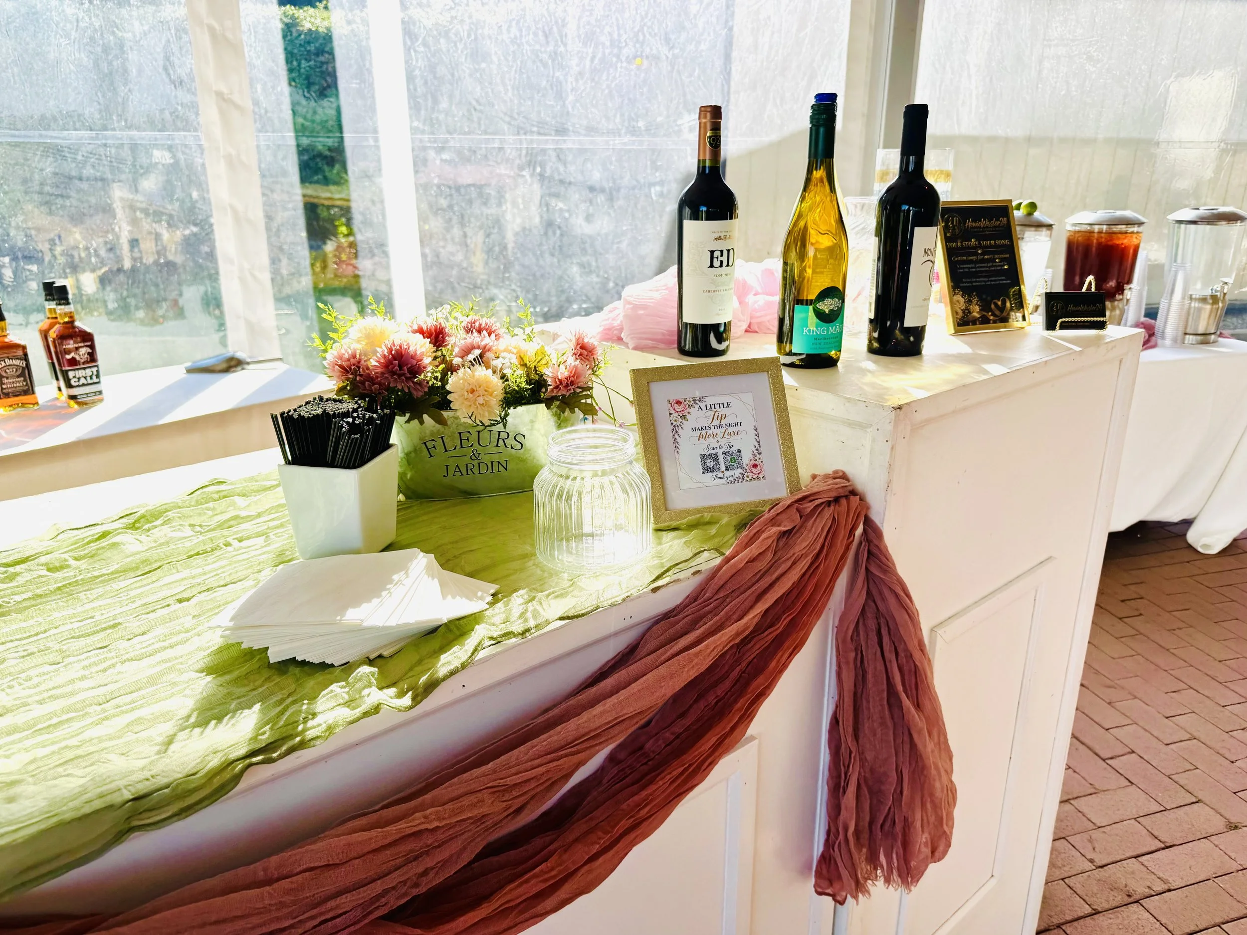 Table decorated with flowers, bottles of wine, and a framed sign, set near a window with sunlight streaming through.