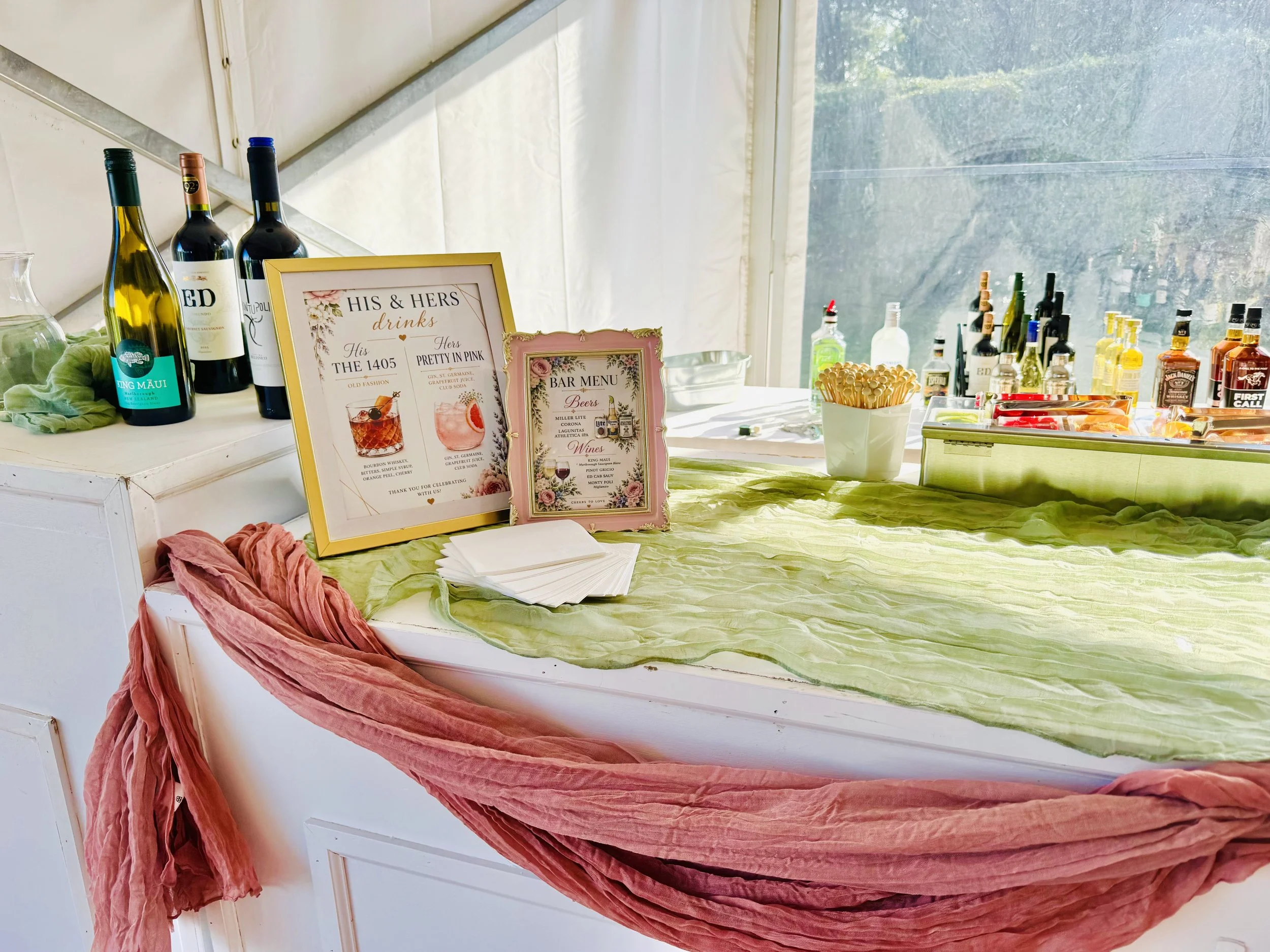 A bar setup with wine bottles, framed signs, and colorful decorations on a table covered with a green cloth.
