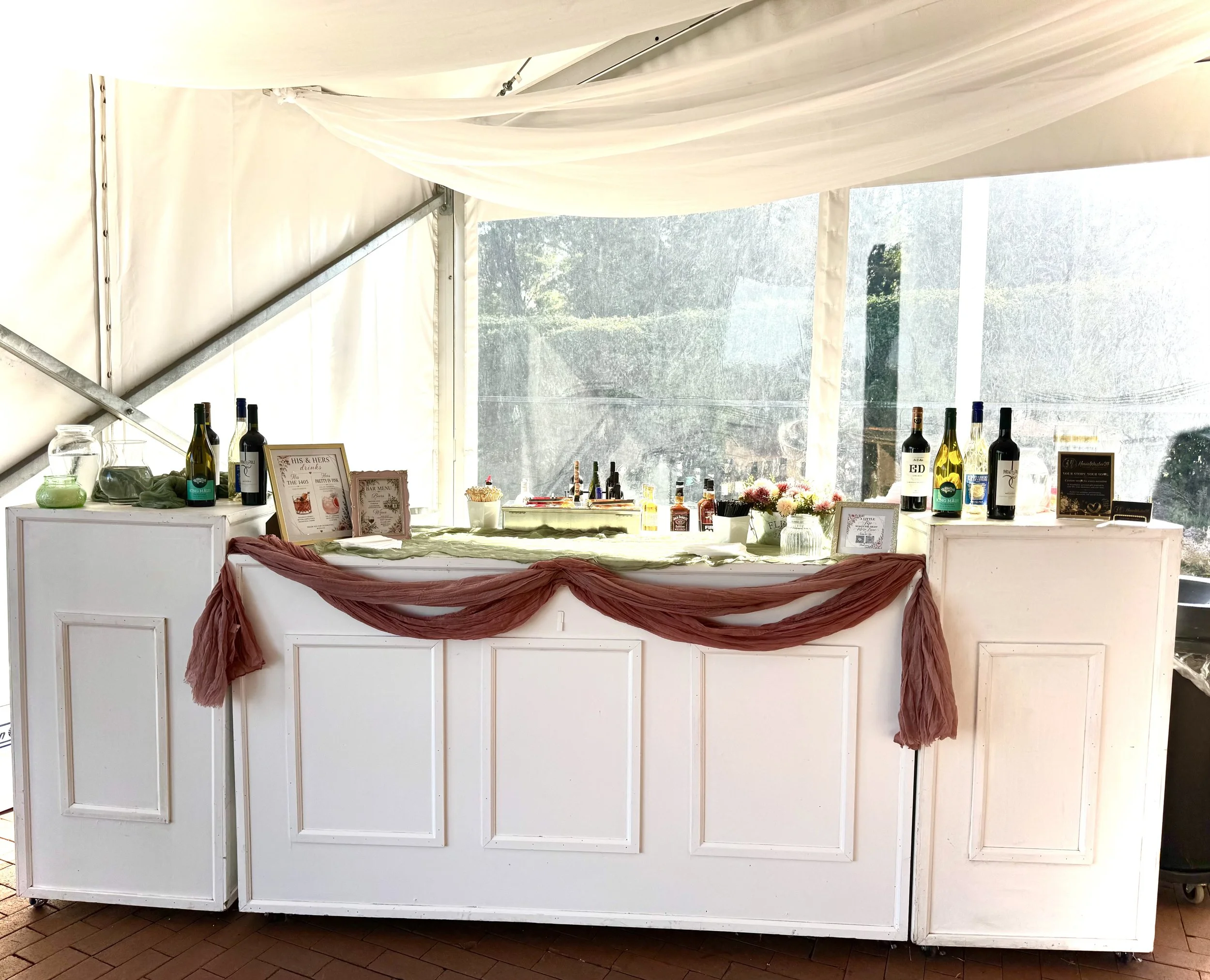 A white bar or counter decorated with a brown cloth draped across the front. On the counter, there are bottles of wine, condiments, flowers, and a small framed menu. The structure appears to be inside a tent with a transparent plastic window revealin