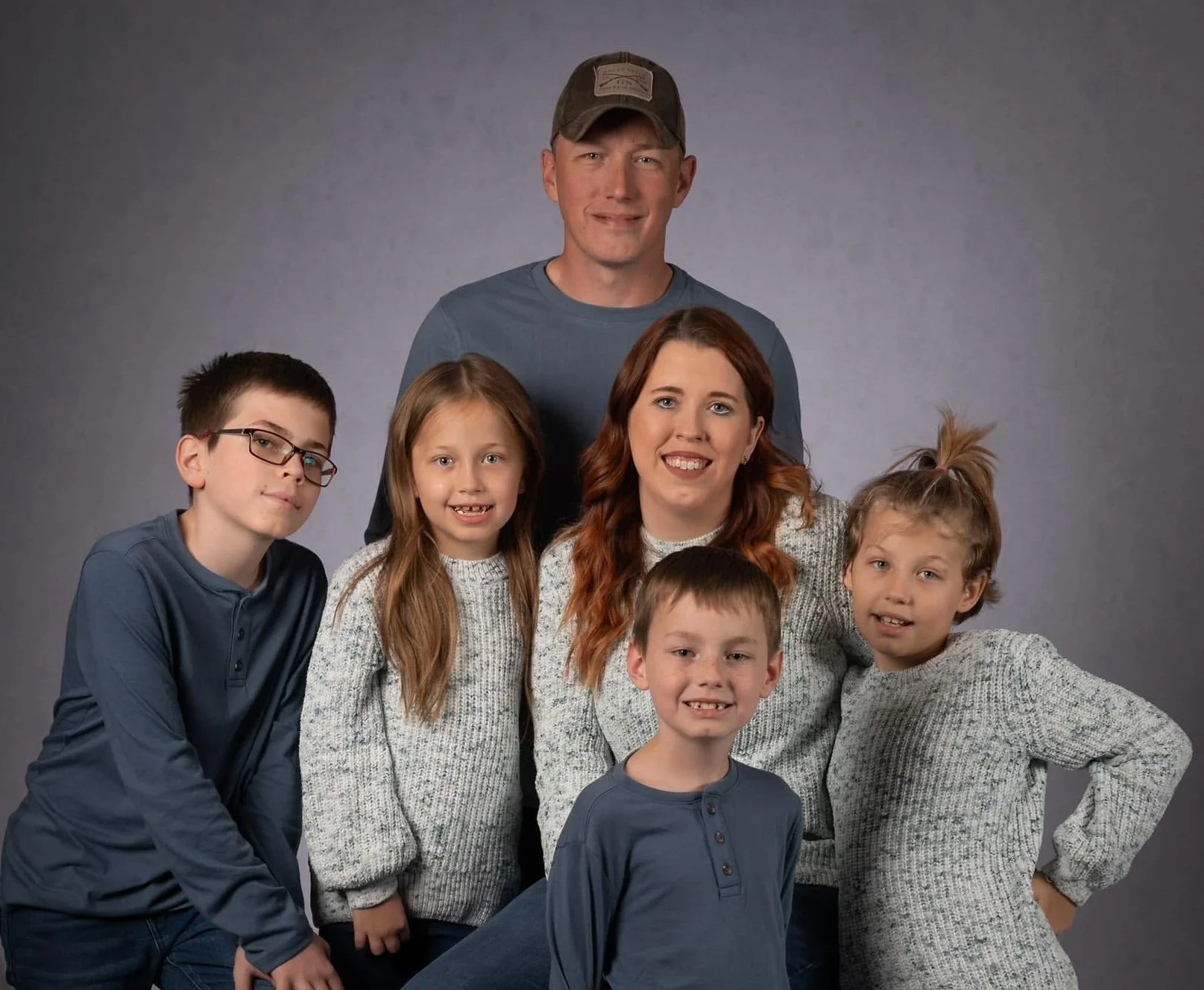 A family portrait of seven people, including five children and two adults, smiling and posing against a gray background.