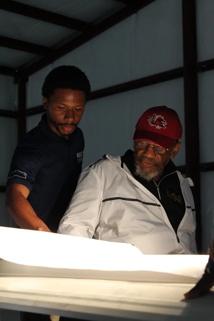 Two men, one younger and one older, looking at a bright object, possibly a document or a tablet, in a dimly lit room. The younger man has dark skin, black curly hair, and wears a dark polo shirt. The older man has dark skin, gray facial hair, glasses, a red baseball cap, and a white jacket with black accents.