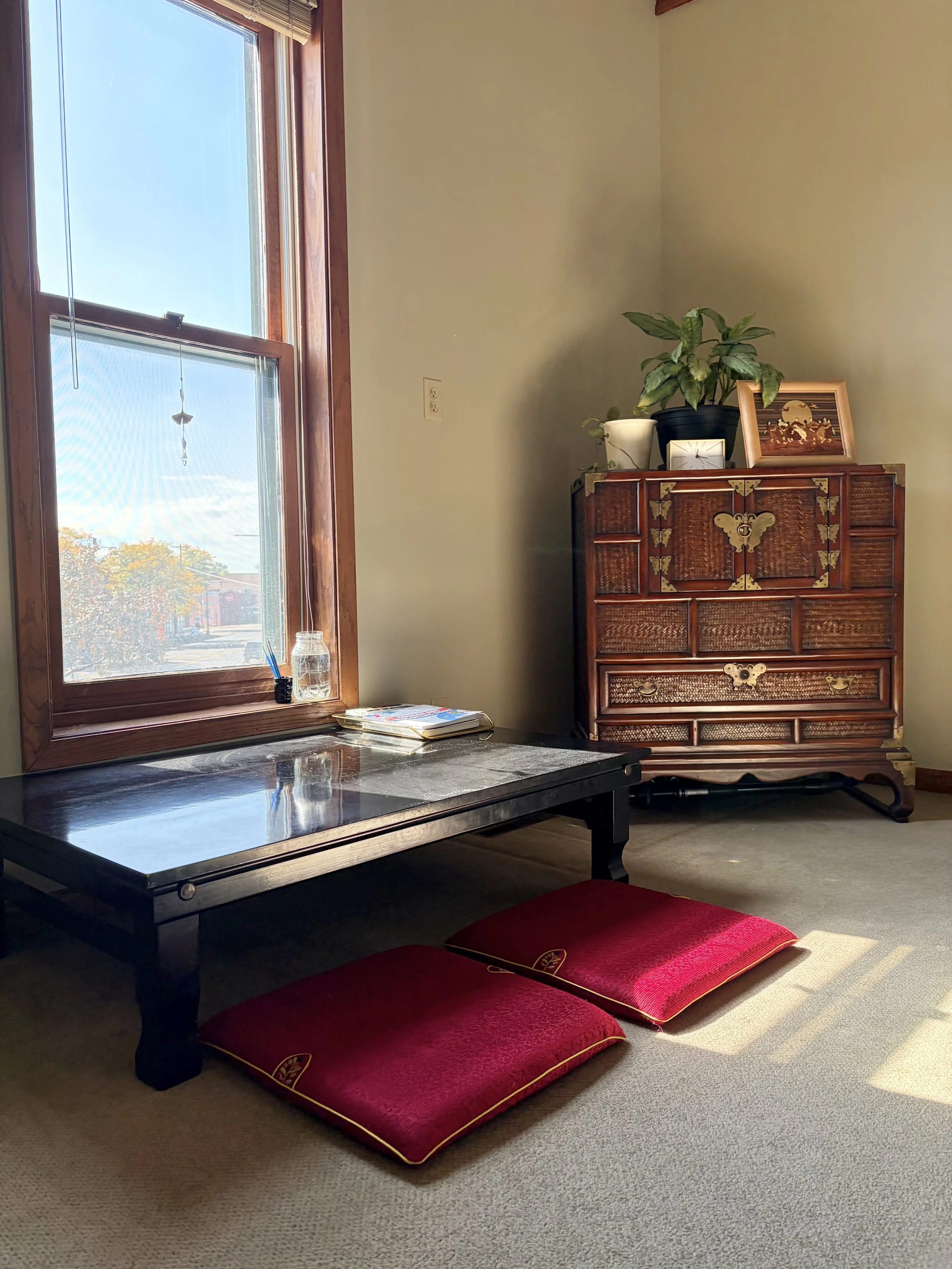 A sunlit corner with a low black lacquer bapsang table and two red floor cushions beside a tall window, and a Korean wooden blanket chest with brass butterfly fittings and a plant on top.