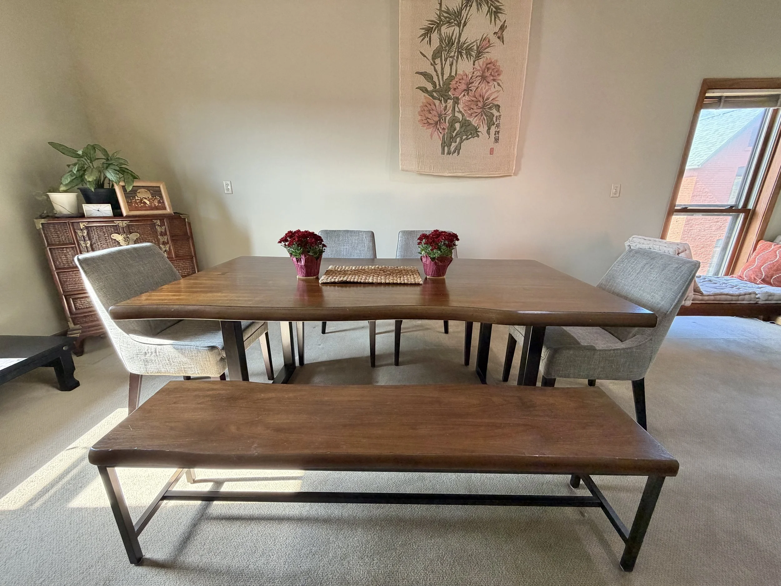 A wooden dining table with four gray upholstered chairs and a long bench, set with two pots of red flowers, in a sunlit room with a floral wall hanging.