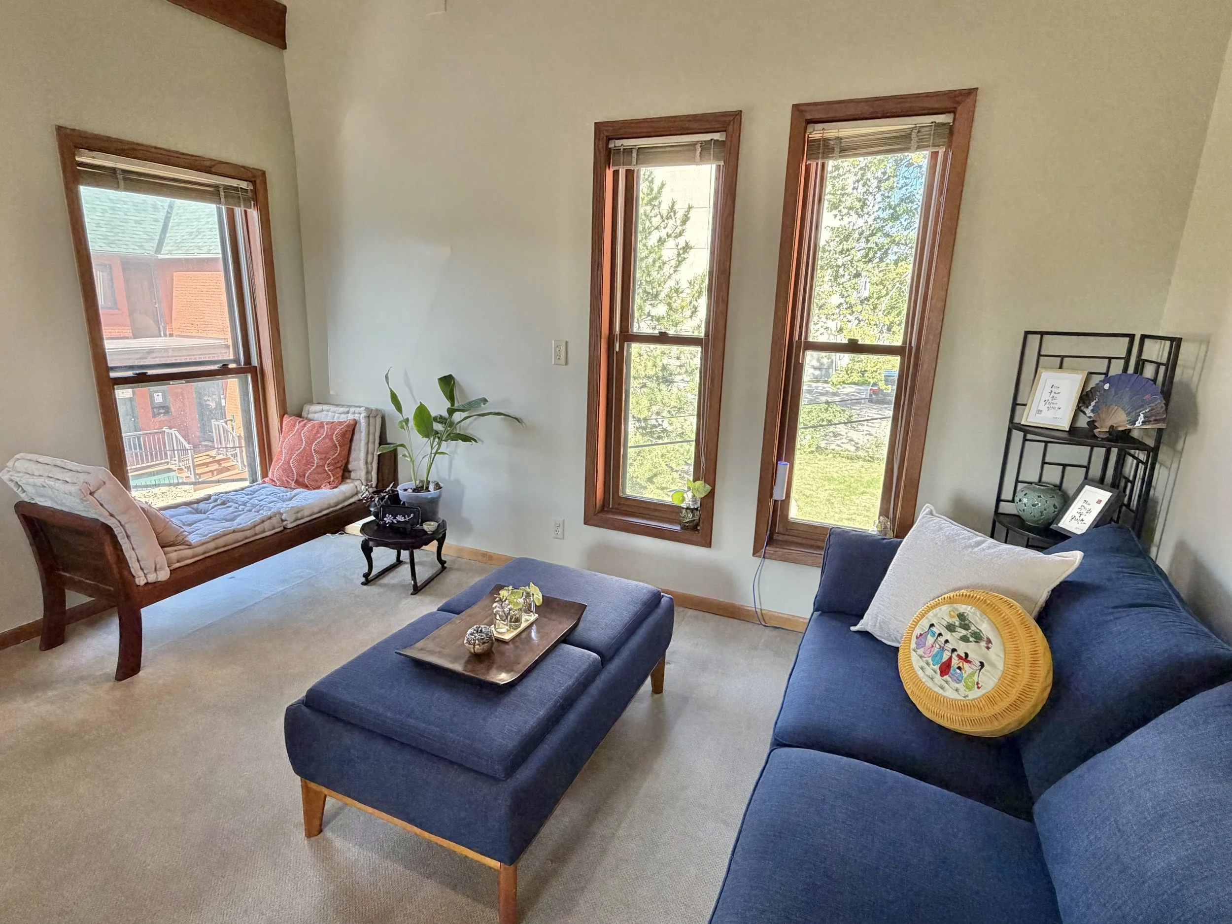 A sunlit room with tall wooden-framed windows, a blue sofa and ottoman, and a cushioned daybed by the window, with plants and framed Korean calligraphy.