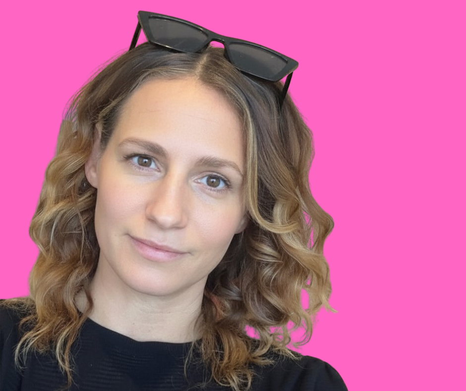 A young woman with curly blonde hair wearing a black top and sunglasses on her head, standing against a pink background.