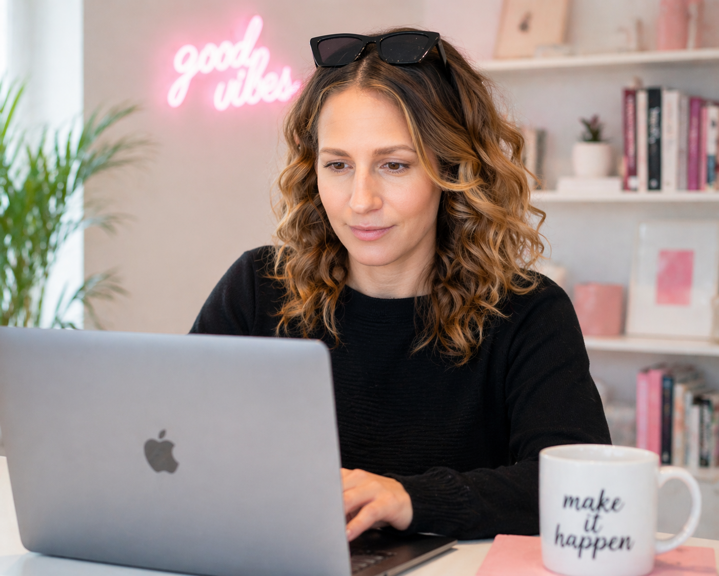 A woman with curly hair and sunglasses on her head is working on a silver MacBook at a desk. There is a white mug with the words 'make it happen' on the desk. In the background, a pink neon sign says 'good vibes' and there are shelves with books and decorative items.