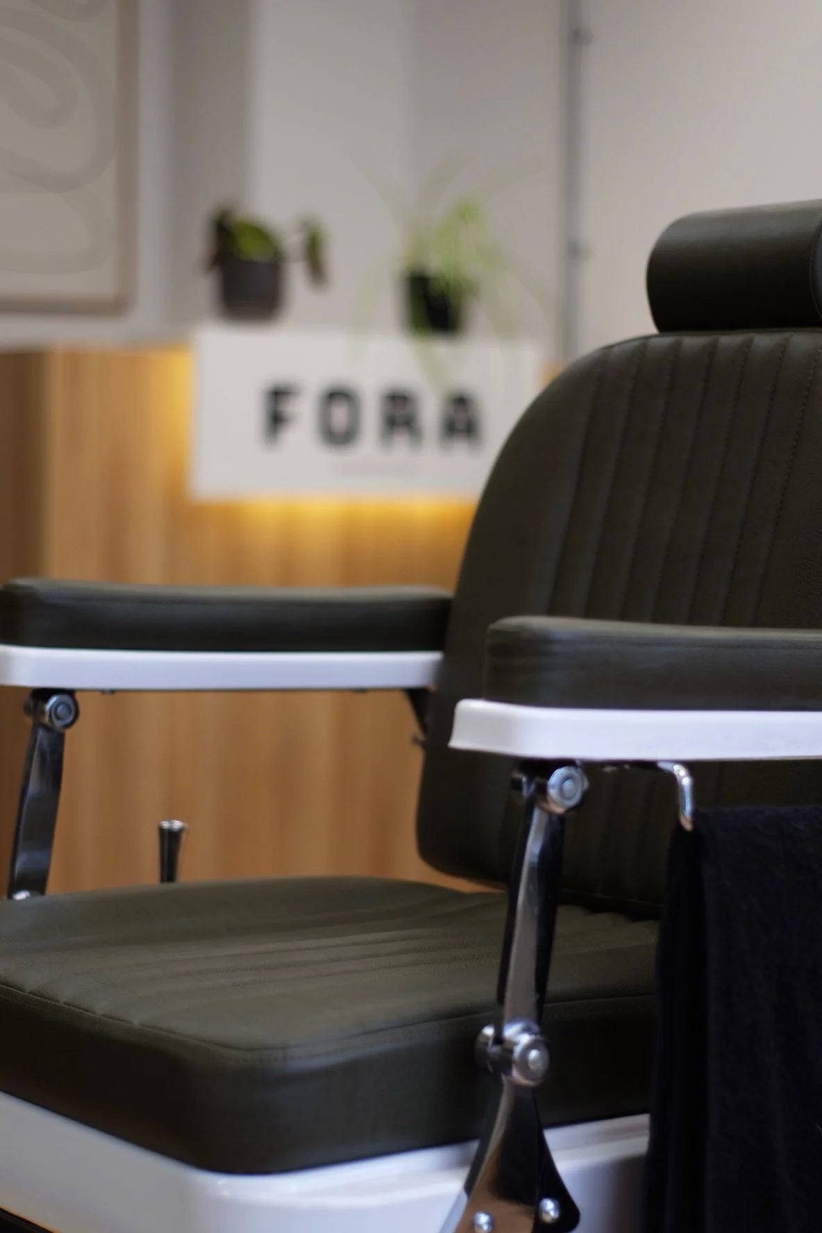 Office waiting room with black chairs and a white armrest, in front of a wooden wall with a blurred white sign that has the letters 'FORA' and potted plants above.