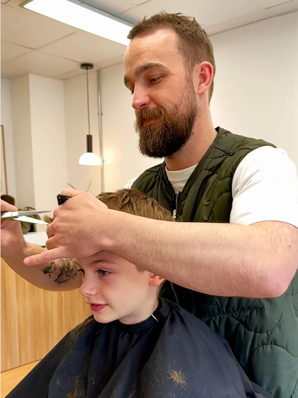 A man with a beard gives a boy a haircut in a salon, with the man holding scissors and the boy covered in a cape.