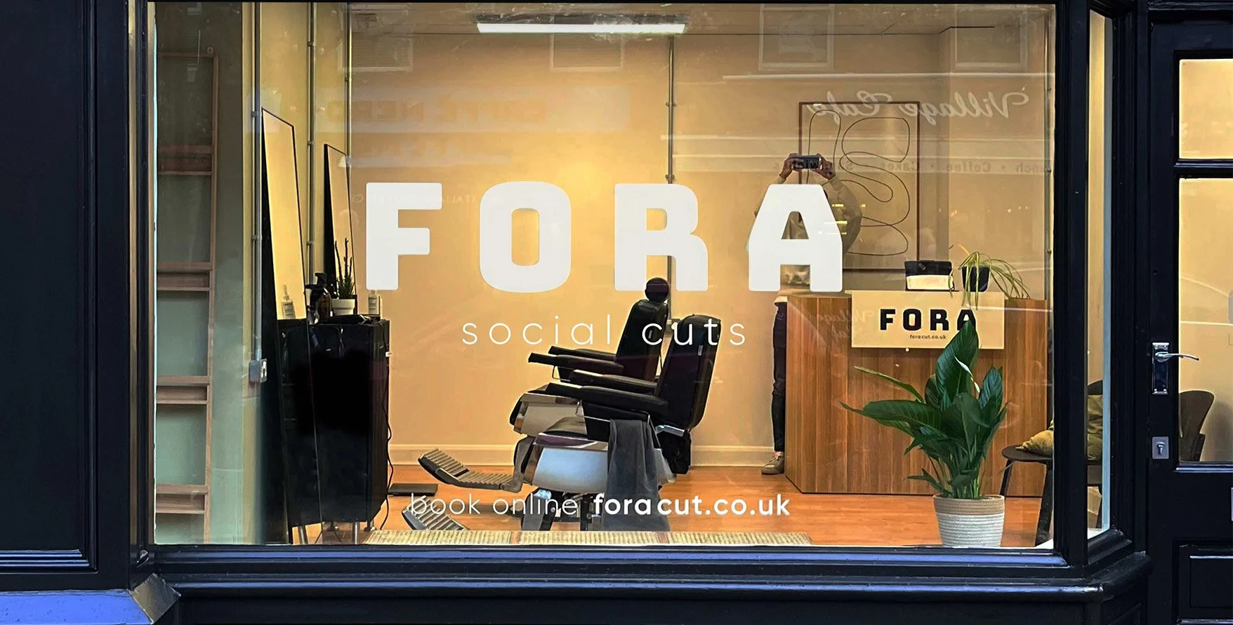 Inside a barber shop with a sign on the window reading 'FORA social cuts' and a message to book online at foracut.co.uk. The space contains barber chairs, a model with mirrors, a wooden reception desk, and a large potted plant.
