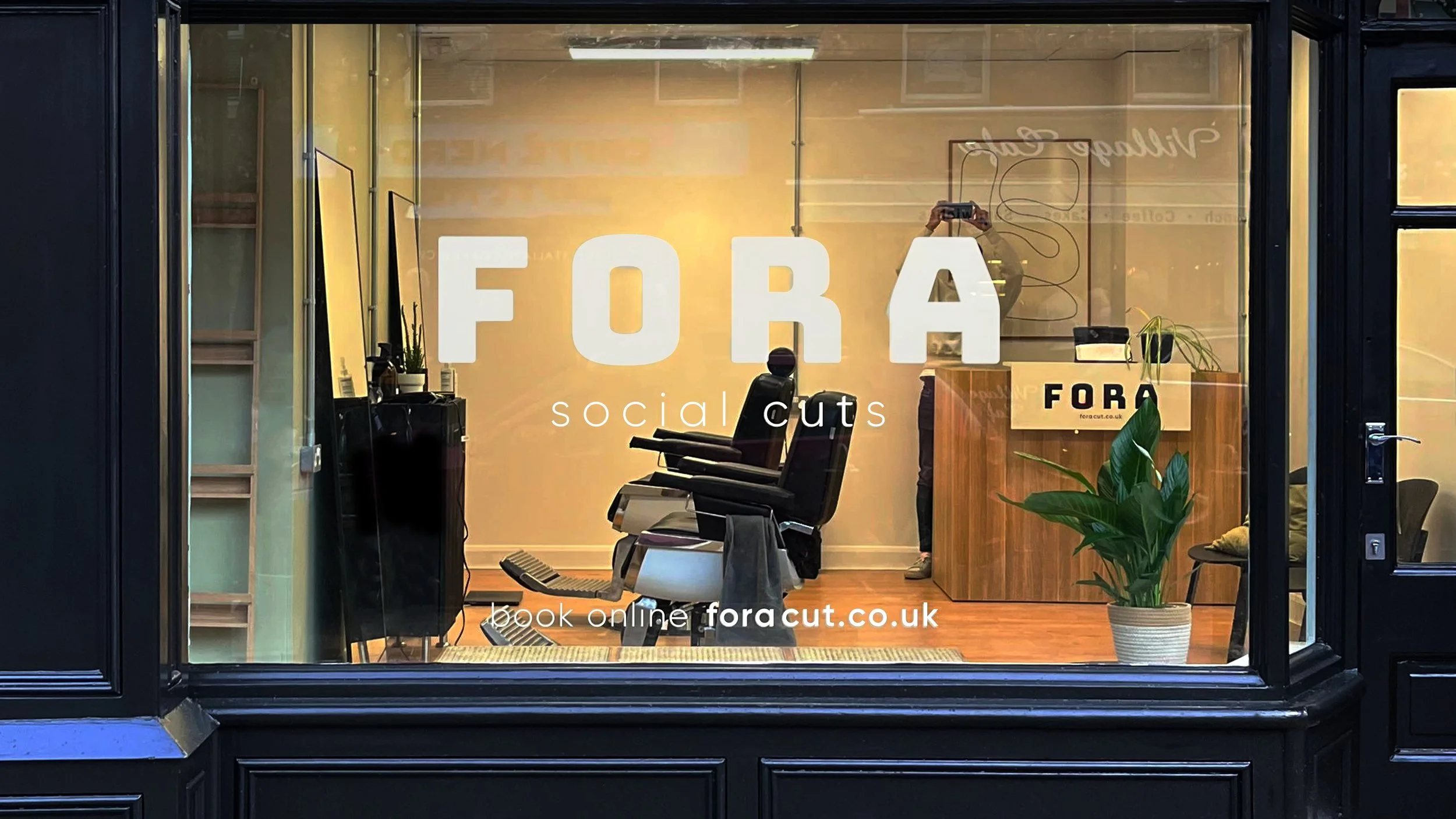 Front window of a salon with the words 'FORA social cuts' and 'book online foracut.co.uk' on the glass, showing interior chairs, a plant, and a person taking a photo with a smartphone.