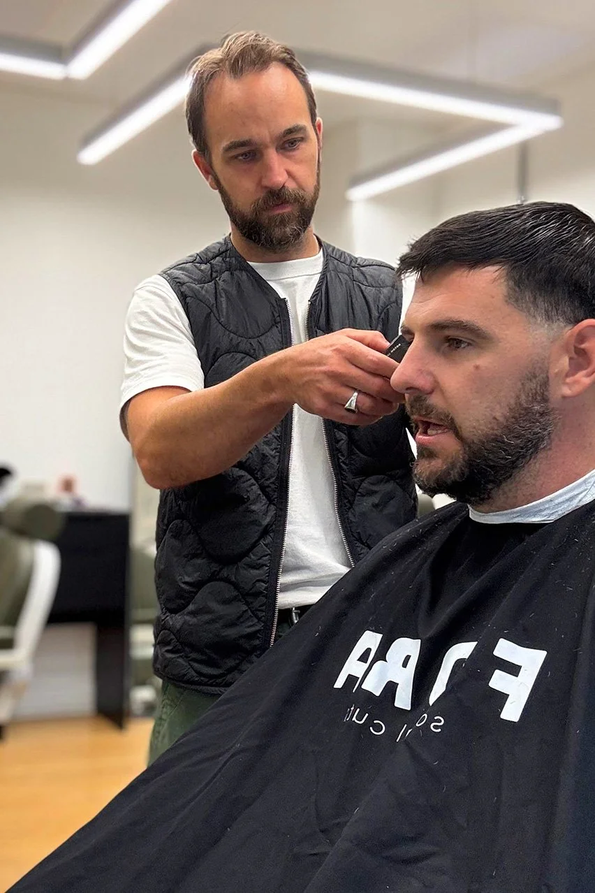 A man getting a haircut at a barbershop, sitting in a barber's chair with a cape, while a barber trims his hair with clippers.