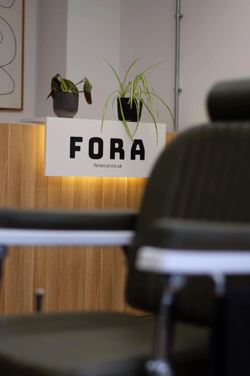 Indoor office reception area with plants on a wooden counter and a sign that says 'FORA' with a website link.