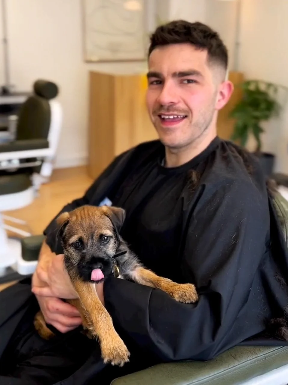 A man with short dark hair and a beard holding a small gray and brown puppy with a pink tongue sticking out. The man is smiling and sitting in a room with a black barber cape on.