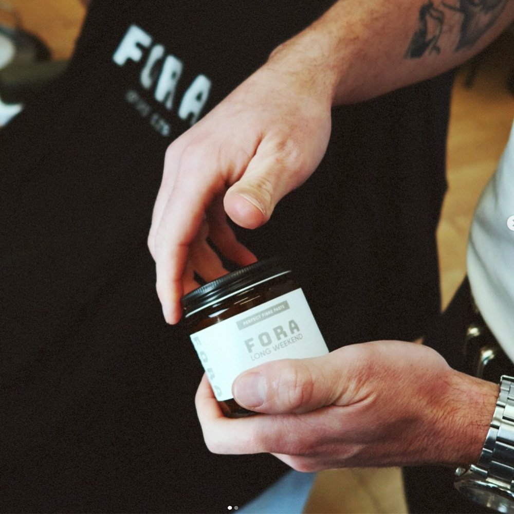 A person holding and opening a small jar labeled 'FORA LONG WEEKEND' with a black lid, wearing a wristwatch and a tattoo on their forearm.