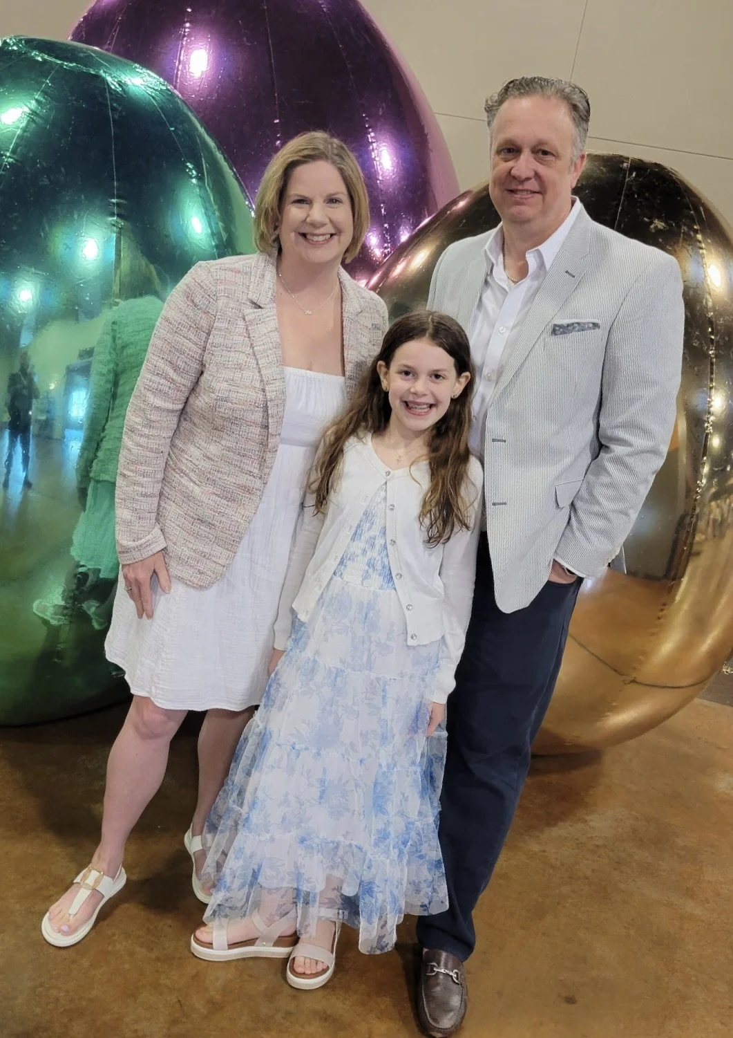 A family of three smiling at the camera at an indoor event with large, shiny, metallic balloons in the background, including a woman, a man, and a young girl.