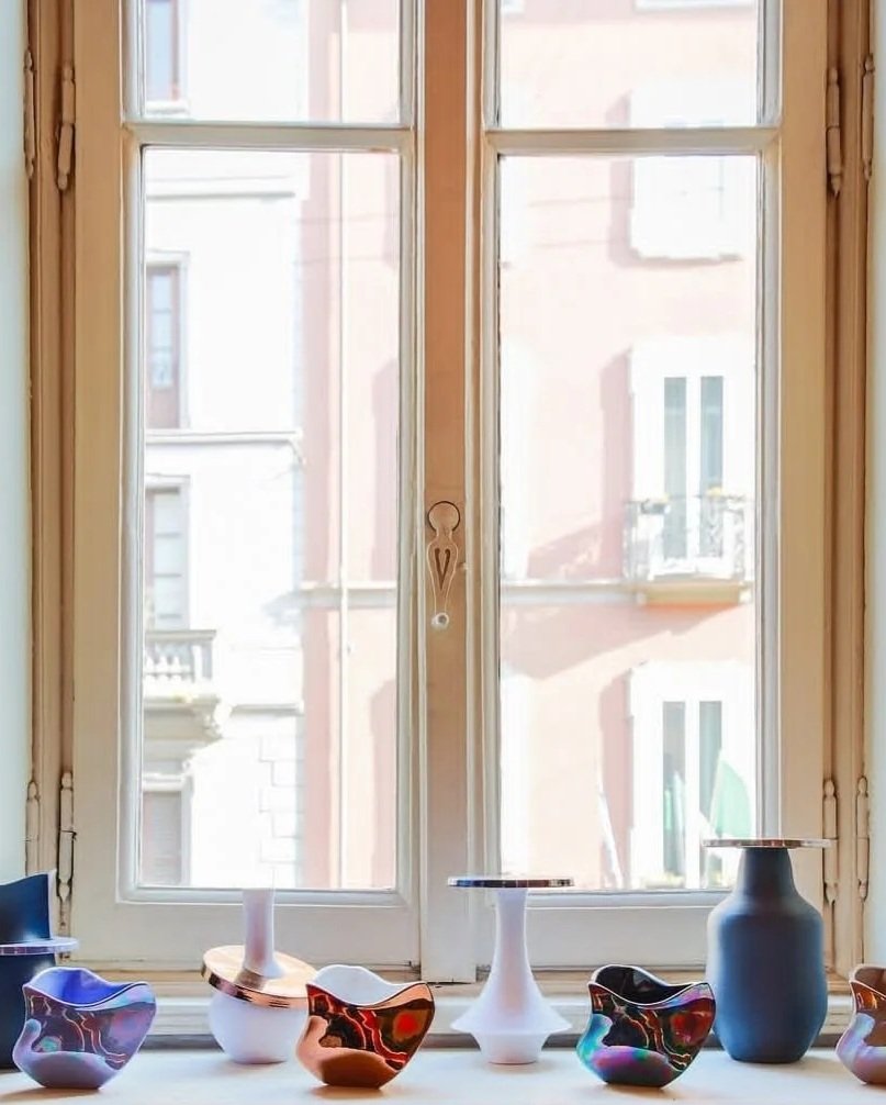 Vasi di vari colori e forme su una finestra, con edifici urbani visibili fuori.