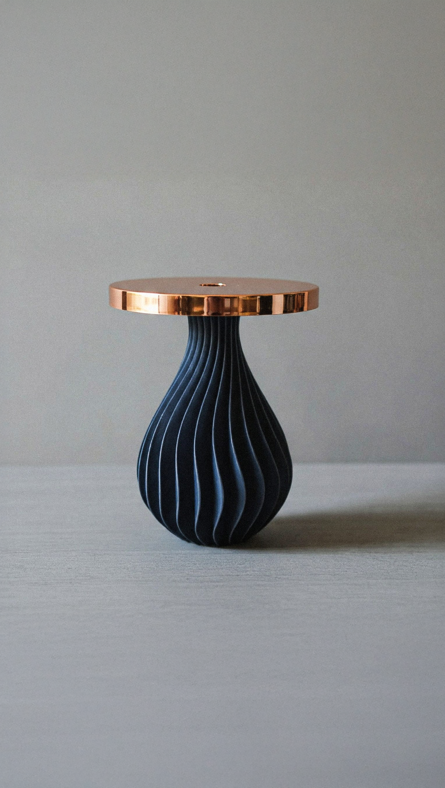 Una tavola con un panno di colore naturale e una figura decorativa con forma di goccia, blu con strisce bianche, e sopra un piatto rotondo di metallo color rame.