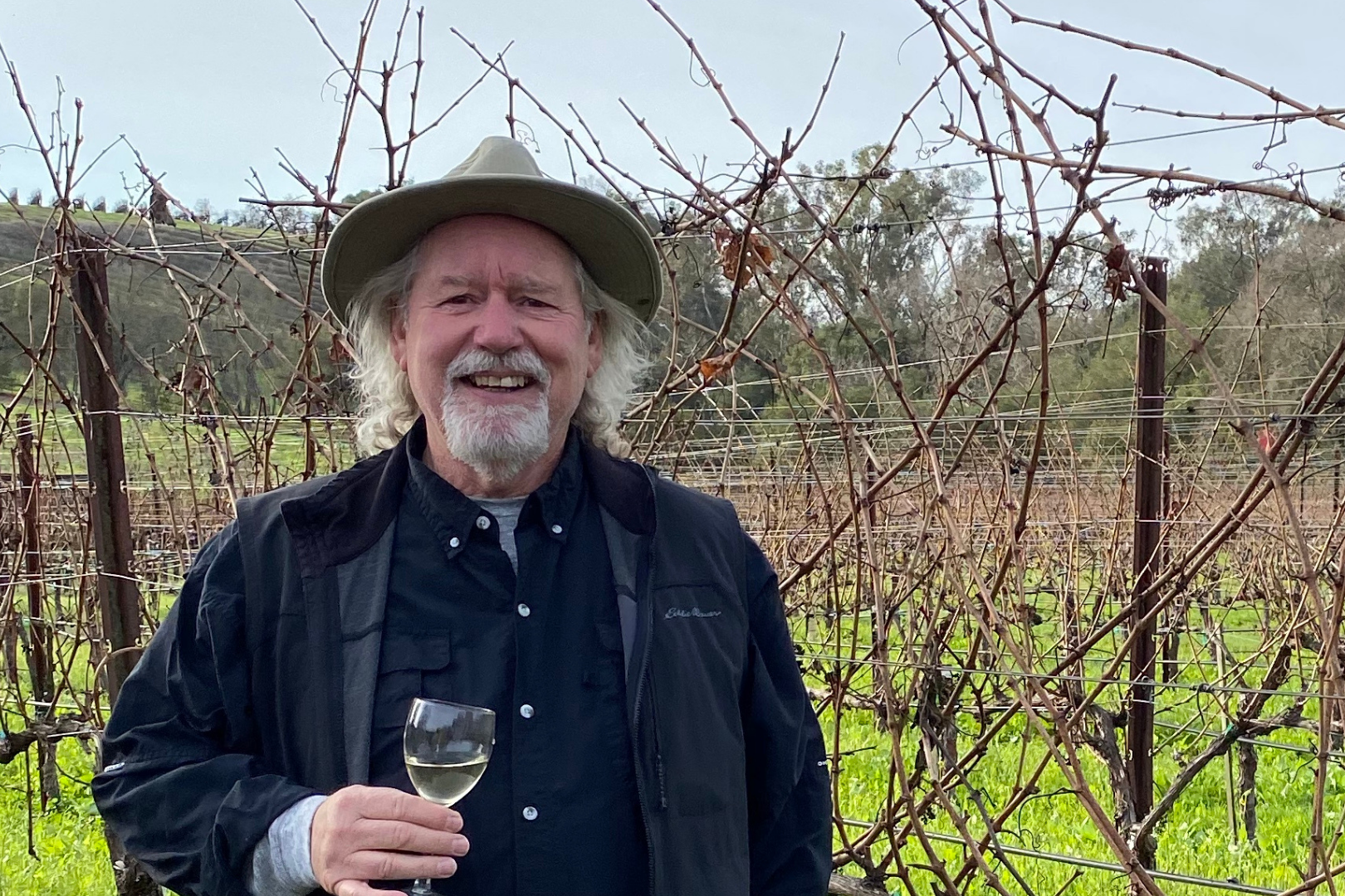 Jim Milone smiling and holding a glass of white wine amidst a vineyard with leafless vines and a rural landscape in the background.