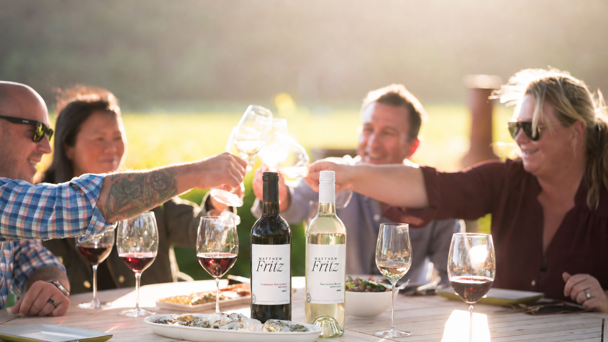 A group of people enjoying a toast at an outdoor dinner party, with wine glasses, bottles, and food on the table.